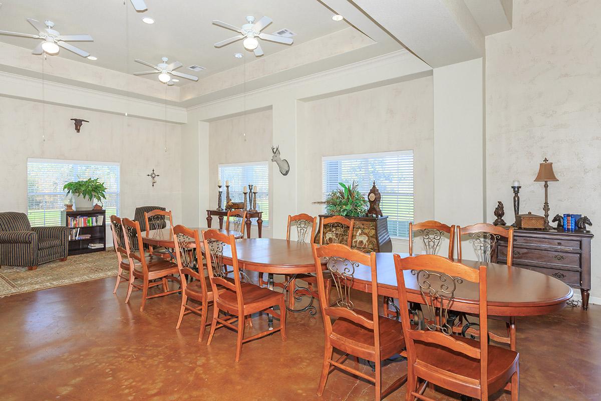 A spacious dining room featuring a large wooden table with multiple chairs, two ceiling fans, and large windows allowing natural light. There are decorative elements including plants, a bookcase, and various decor items on side tables, creating a warm and inviting atmosphere.
