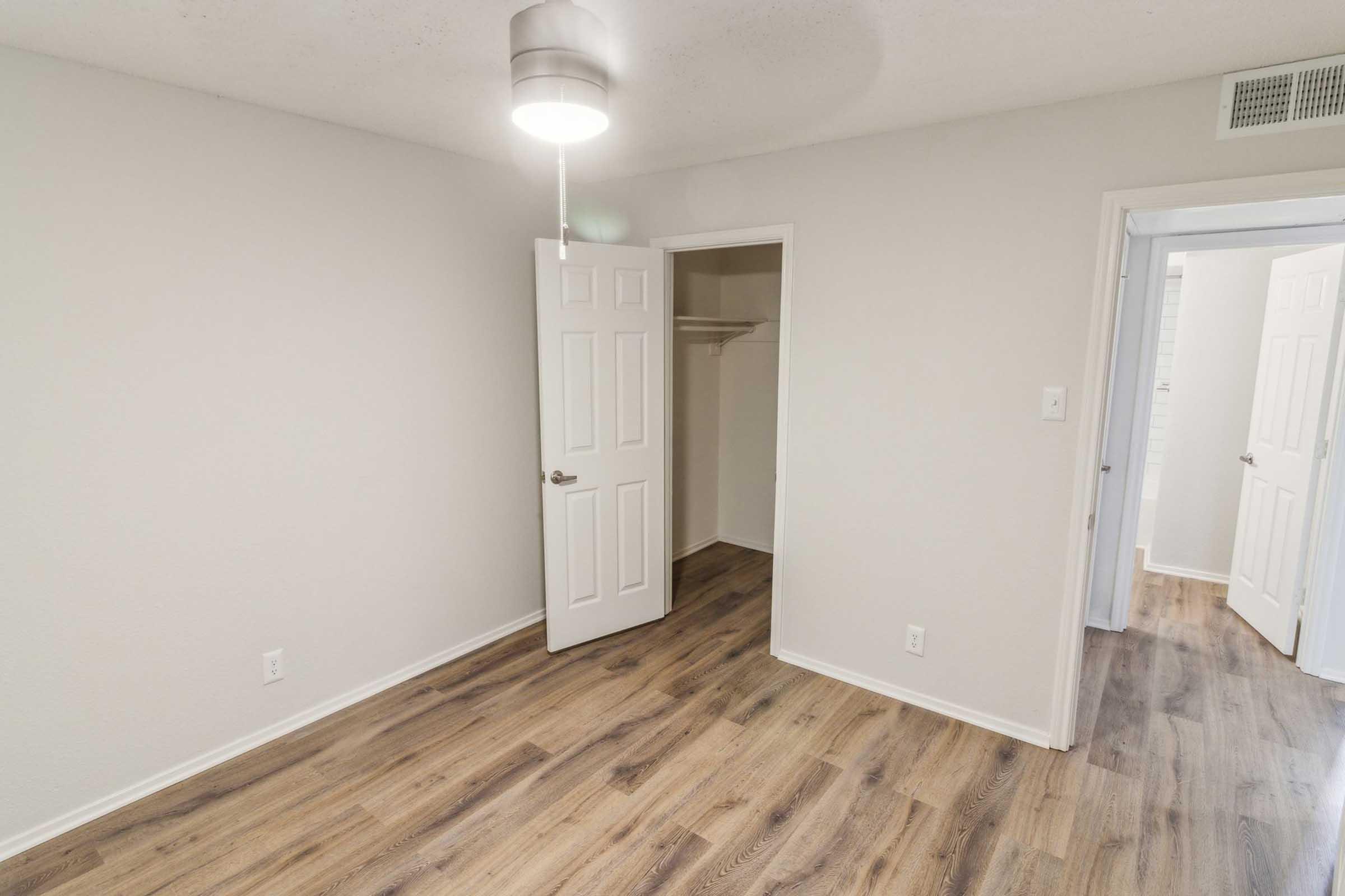 A brightly lit, empty room featuring light-colored walls and a wooden floor. There are two doorways: one leading to a closet and another to an adjacent space. A ceiling light fixture hangs above, creating a welcoming atmosphere. The room appears spacious and well-maintained.
