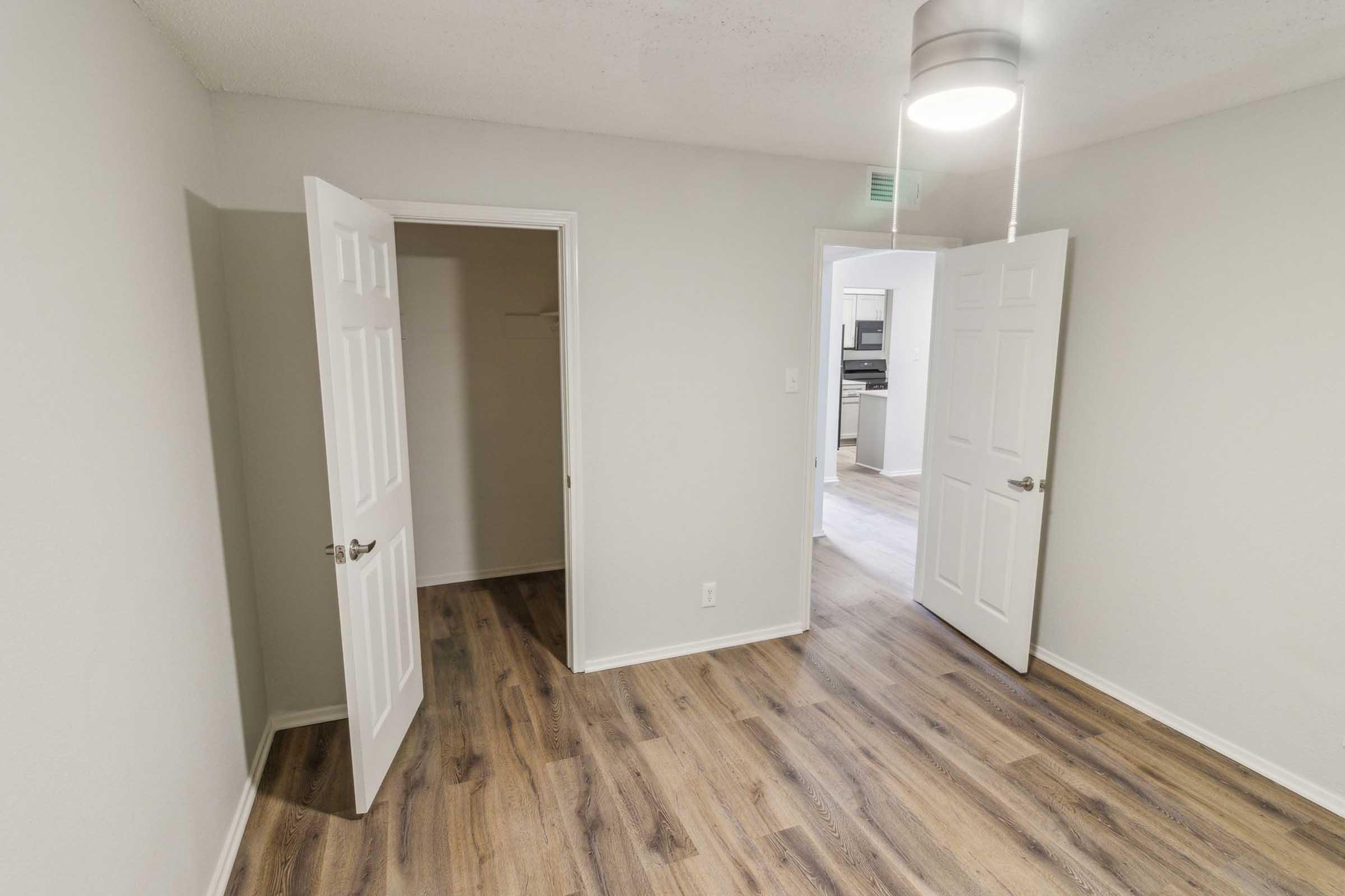 A bright, empty room with light gray walls and laminate flooring. Two white doors are open, one leading to a closet and the other to an adjoining space. A ceiling light fixture hangs above, illuminating the room. The overall atmosphere is clean and modern.