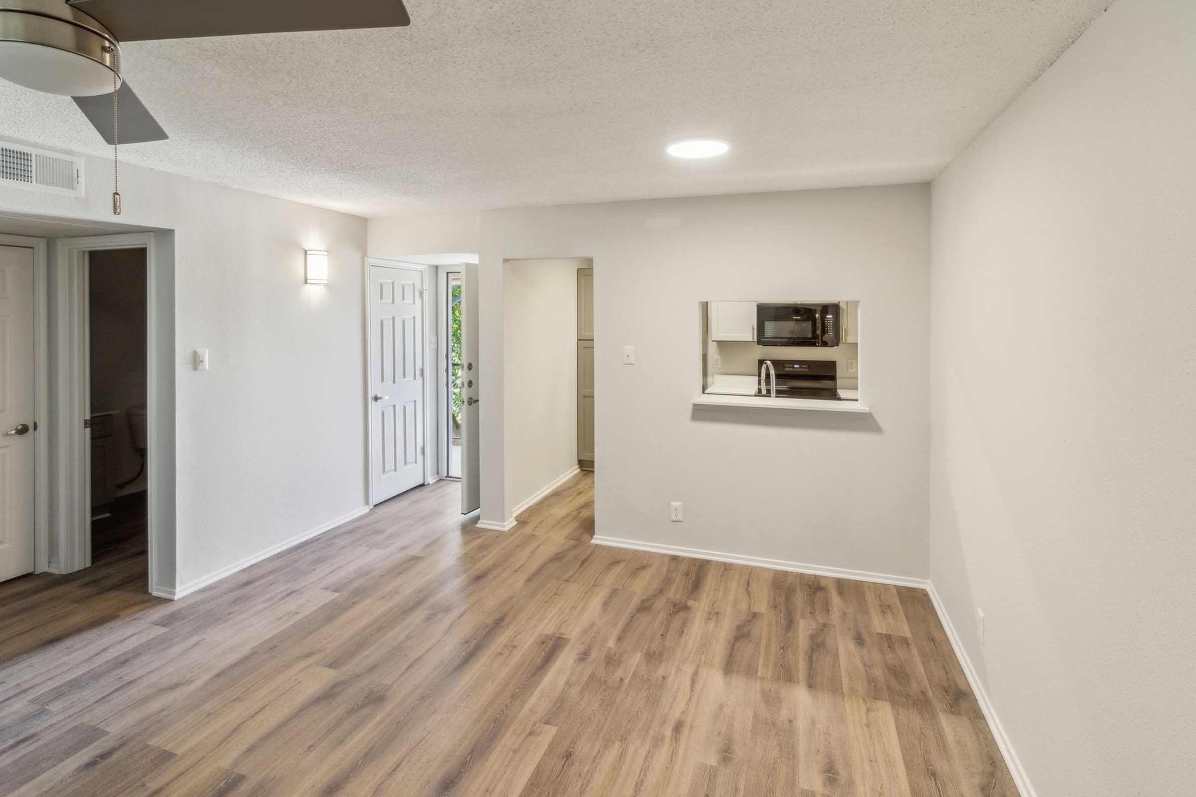 A bright and spacious room with light-colored walls and wood-like flooring. There is a ceiling fan, and a doorway leading to another room. A small opening in the wall reveals a kitchenette area with a sink and a microwave. Natural light illuminates the space, creating a welcoming atmosphere.