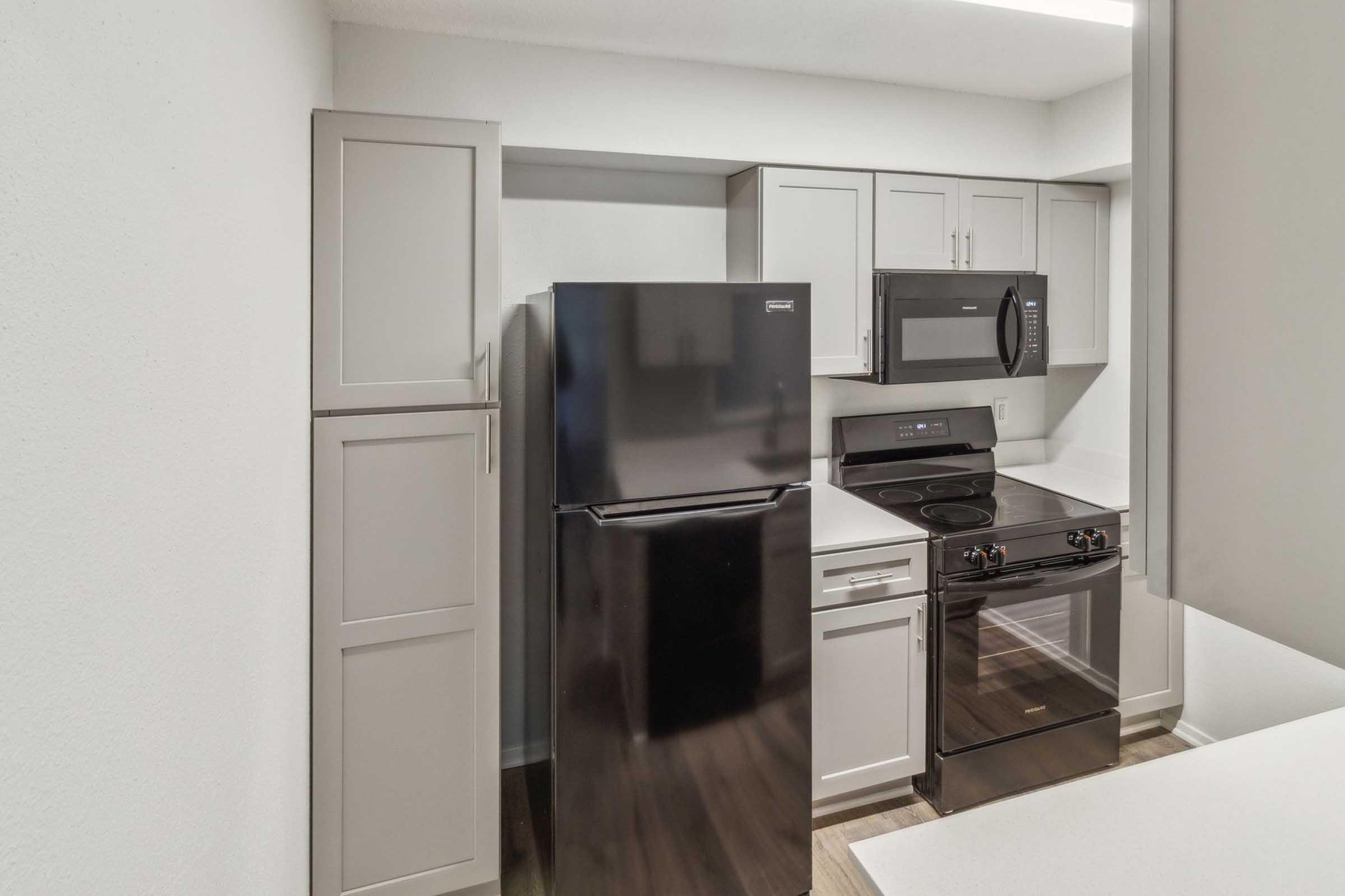 A modern kitchen featuring gray cabinetry, a black refrigerator, a black microwave above an electric stove, and a clean countertop. The space is well-lit, with a minimalist design and wood-like flooring.