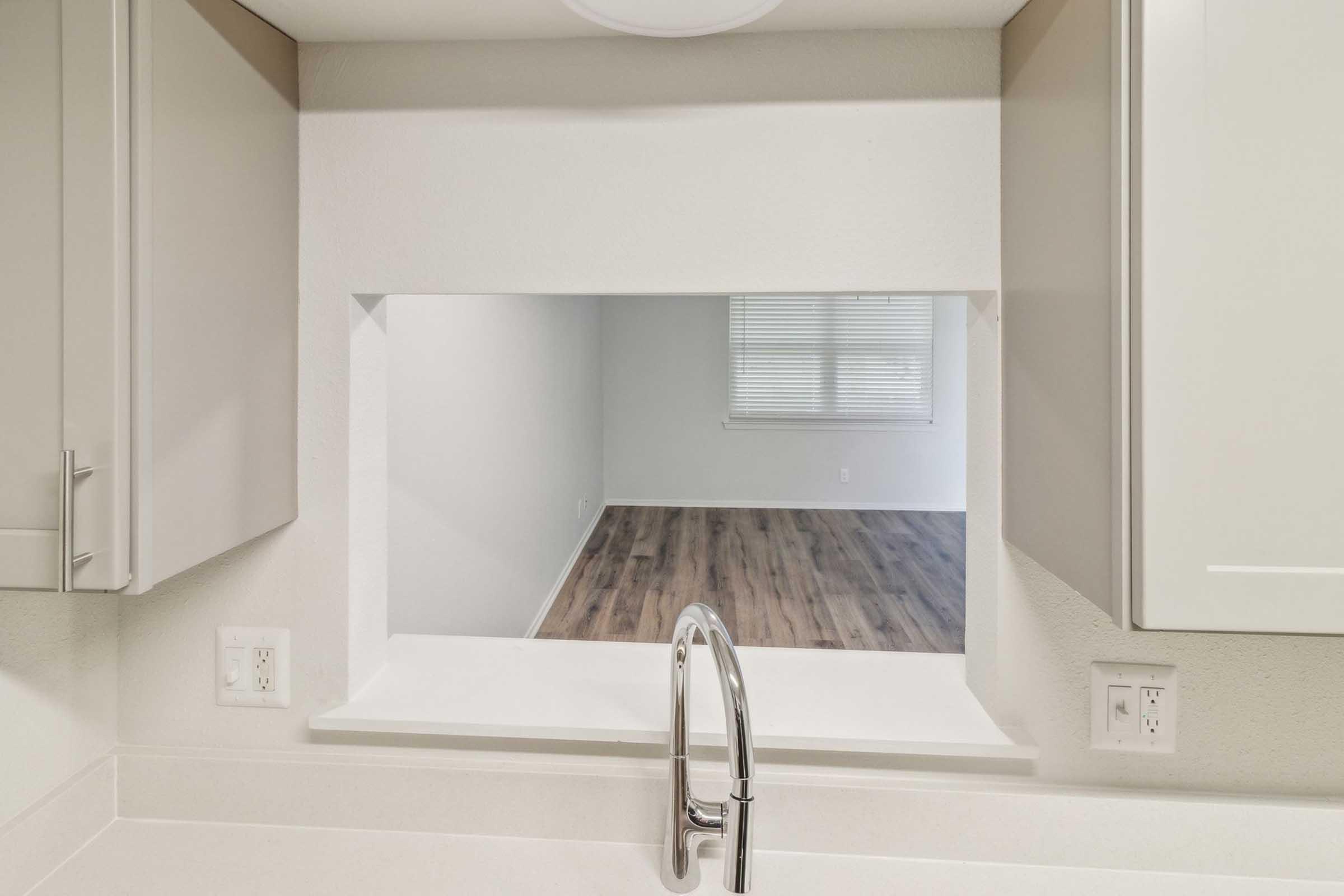 A kitchen view featuring a rectangular pass-through window. The window opens to a spacious, empty living area with a light-colored wall and large windows allowing natural light. The kitchen has modern cabinetry and a sleek faucet in the foreground. The flooring in the living area is wood-like.