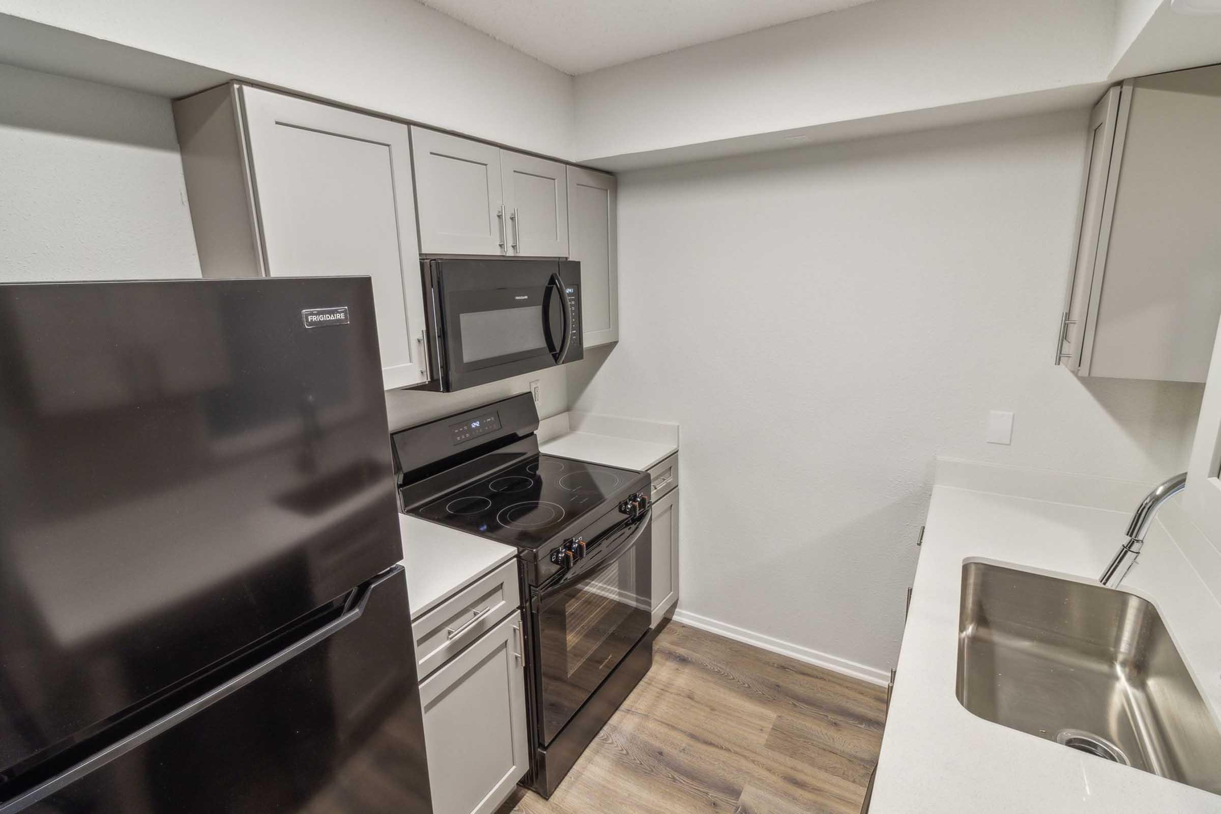 Modern kitchen with gray cabinetry, a black refrigerator, an electric stove, and a microwave above it. The countertop is light-colored, and there's a stainless steel sink. The flooring is wood-like, and the walls are painted light gray.