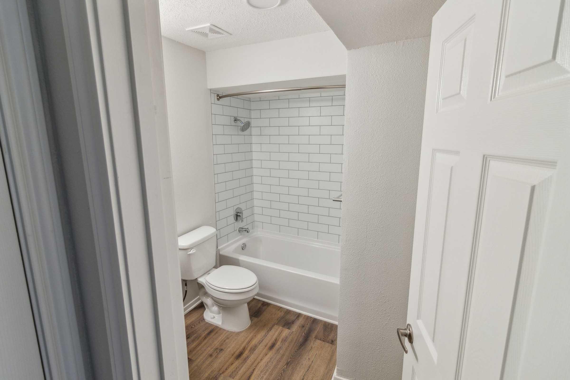 A clean, modern bathroom featuring a toilet, bathtub with a shower, and white subway tile on the walls. The floor is made of light wood, and the space is well-lit with a neutral color scheme. A door partially opens into the room, providing a glimpse of the layout.