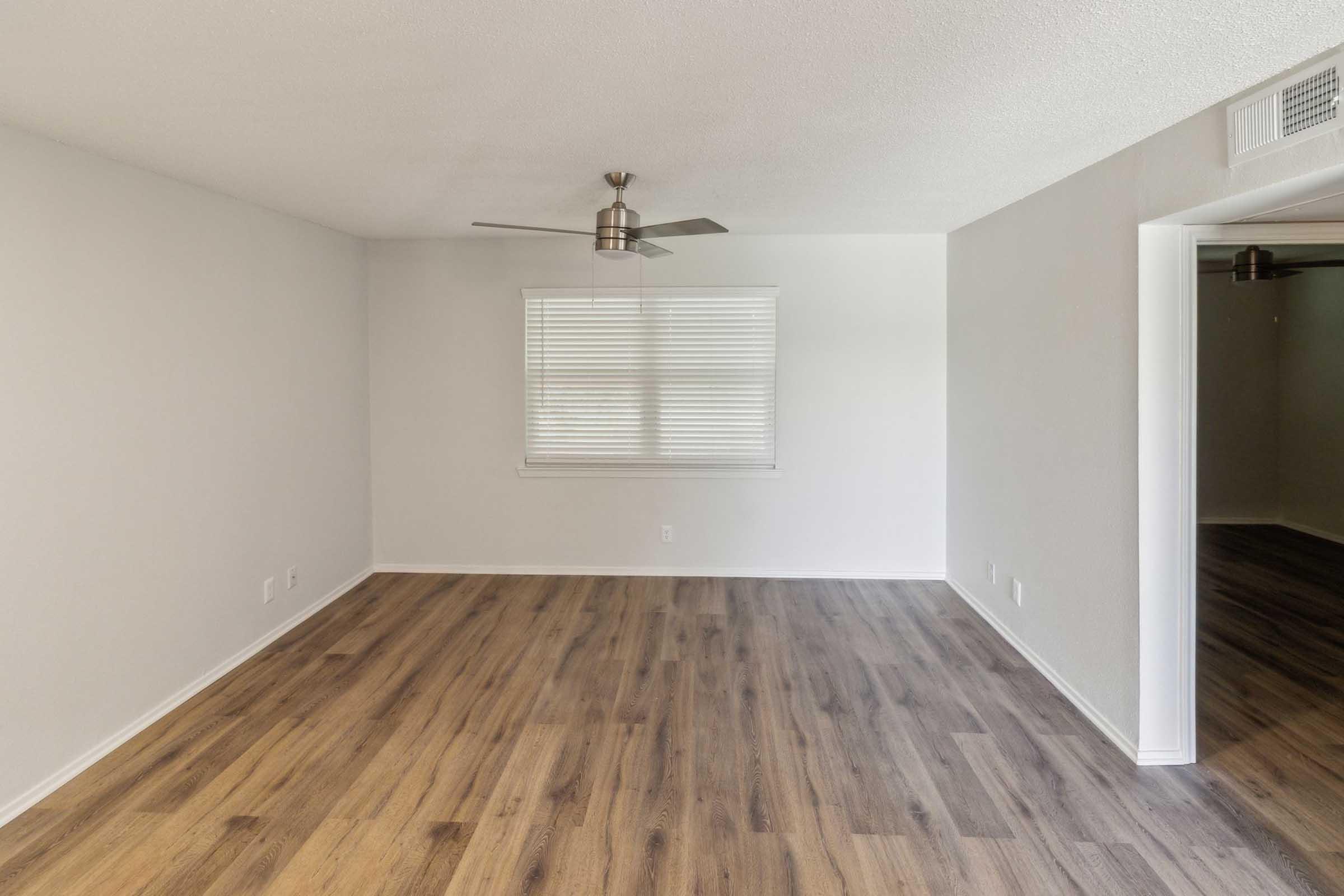 Empty living room featuring light-colored walls, laminate flooring, and a ceiling fan. A window with horizontal blinds allows natural light to enter. The space is bright and minimalistic, creating an open feel. There is a doorway leading to another room visible in the background.