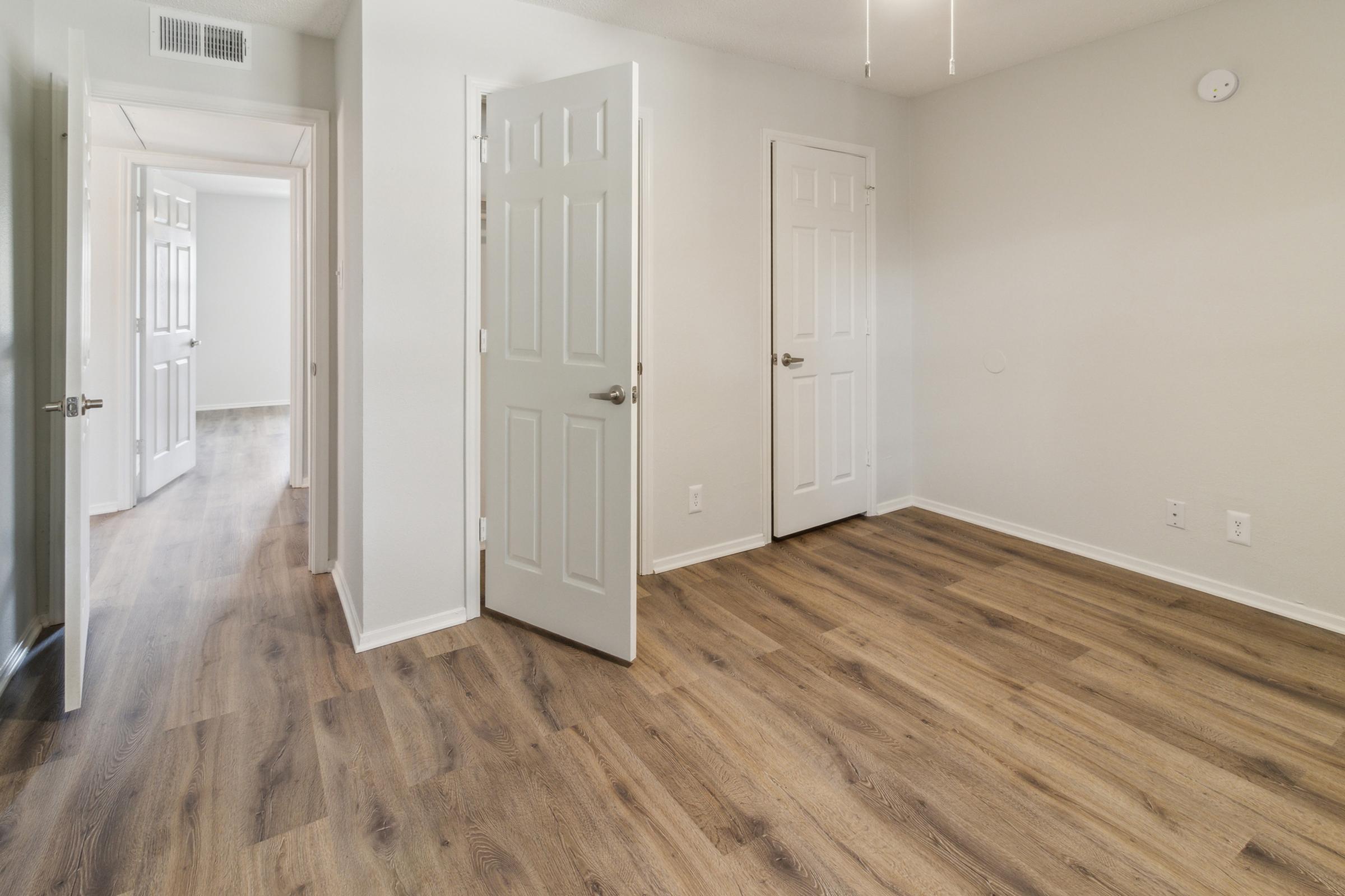 Empty room with light-colored walls and wooden flooring. Two doors on the left lead to adjacent spaces, while a third door is visible on the right. The room is well-lit and appears spacious, featuring a light fixture on the ceiling and a blank wall with no decor.