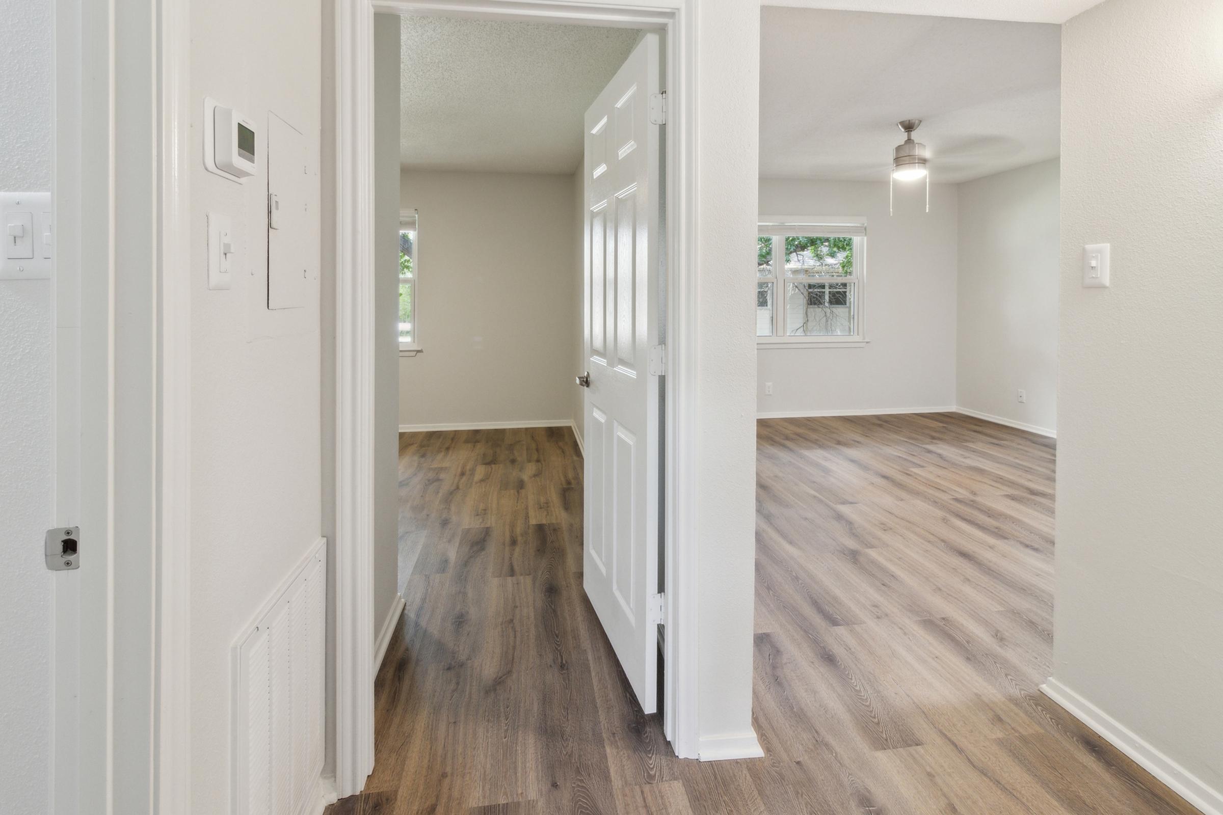A view from a doorway into a modern, light-filled living space with laminate flooring. The room features a ceiling fan, windows, and a neutral color palette, conveying a clean and inviting atmosphere.