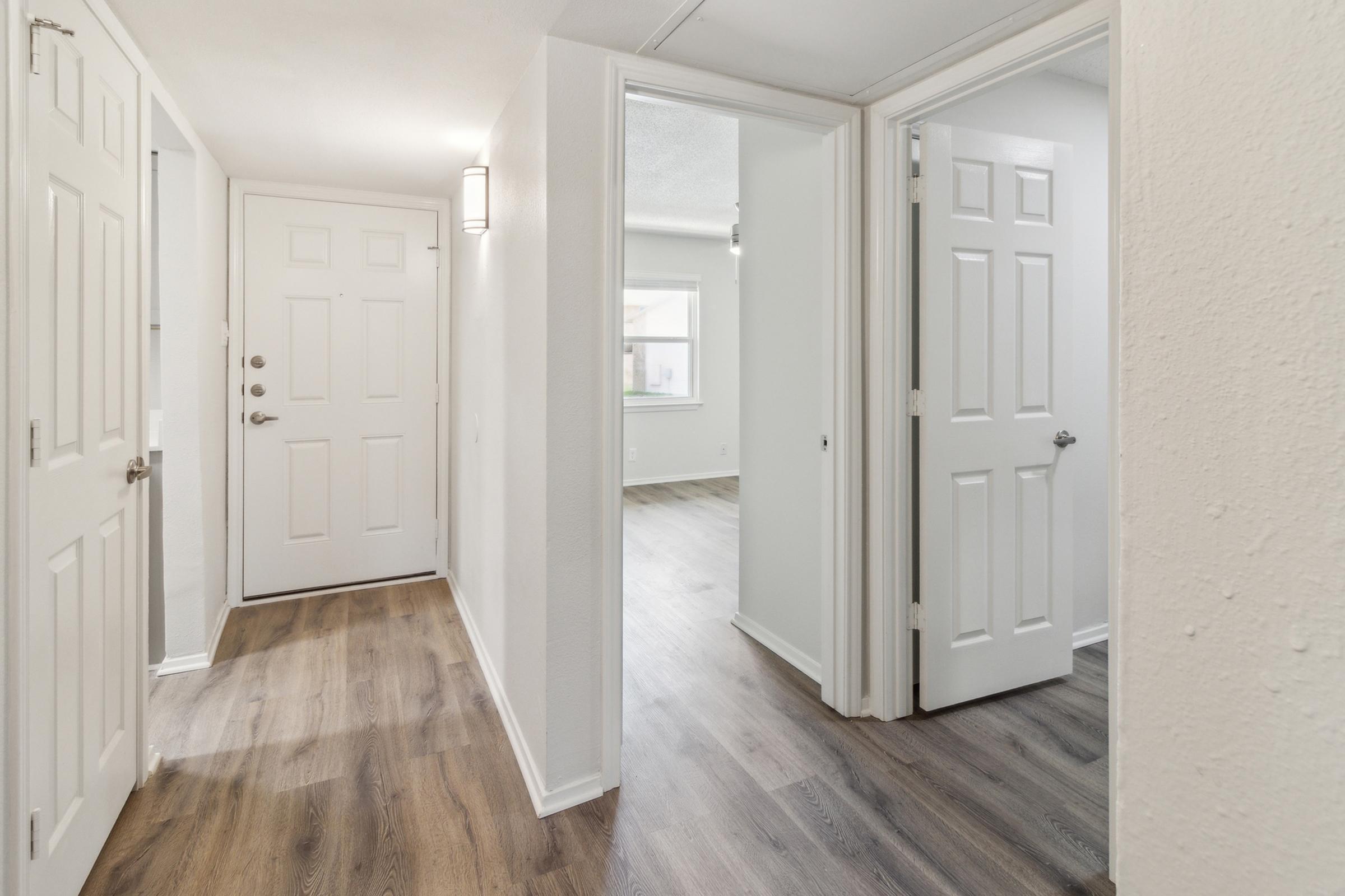 A bright hallway featuring white walls and light wood flooring. There are multiple doors leading to different rooms, with one door slightly ajar, revealing a glimpse of another room. The hallway is well-lit with a wall sconce, creating an inviting atmosphere.