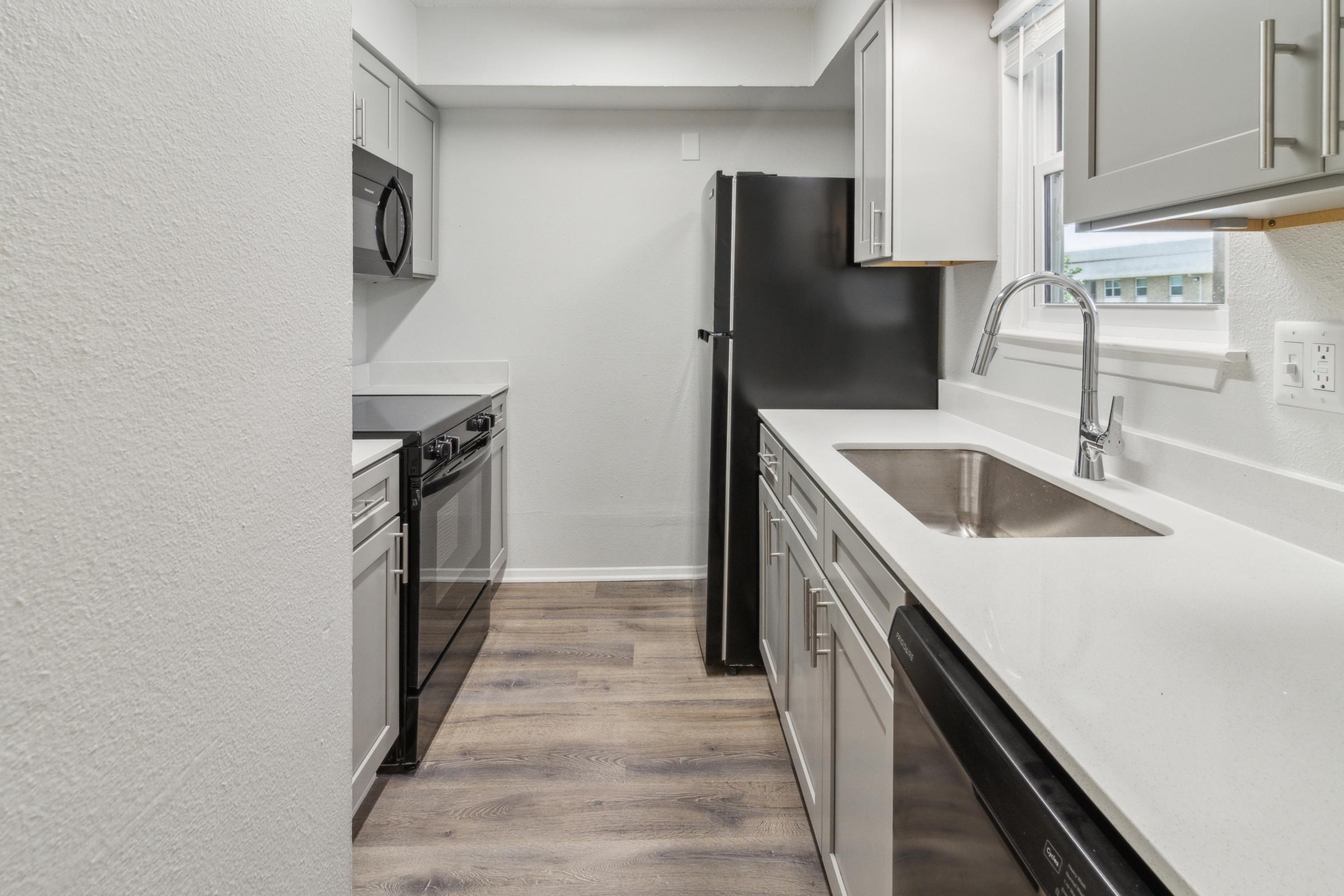 Modern kitchen featuring sleek gray cabinets, stainless steel appliances, including a microwave, dishwasher, and refrigerator. The countertop is white with a stainless steel sink. The floor is timber-style laminate, and natural light flows in from a window, creating a bright and inviting atmosphere.