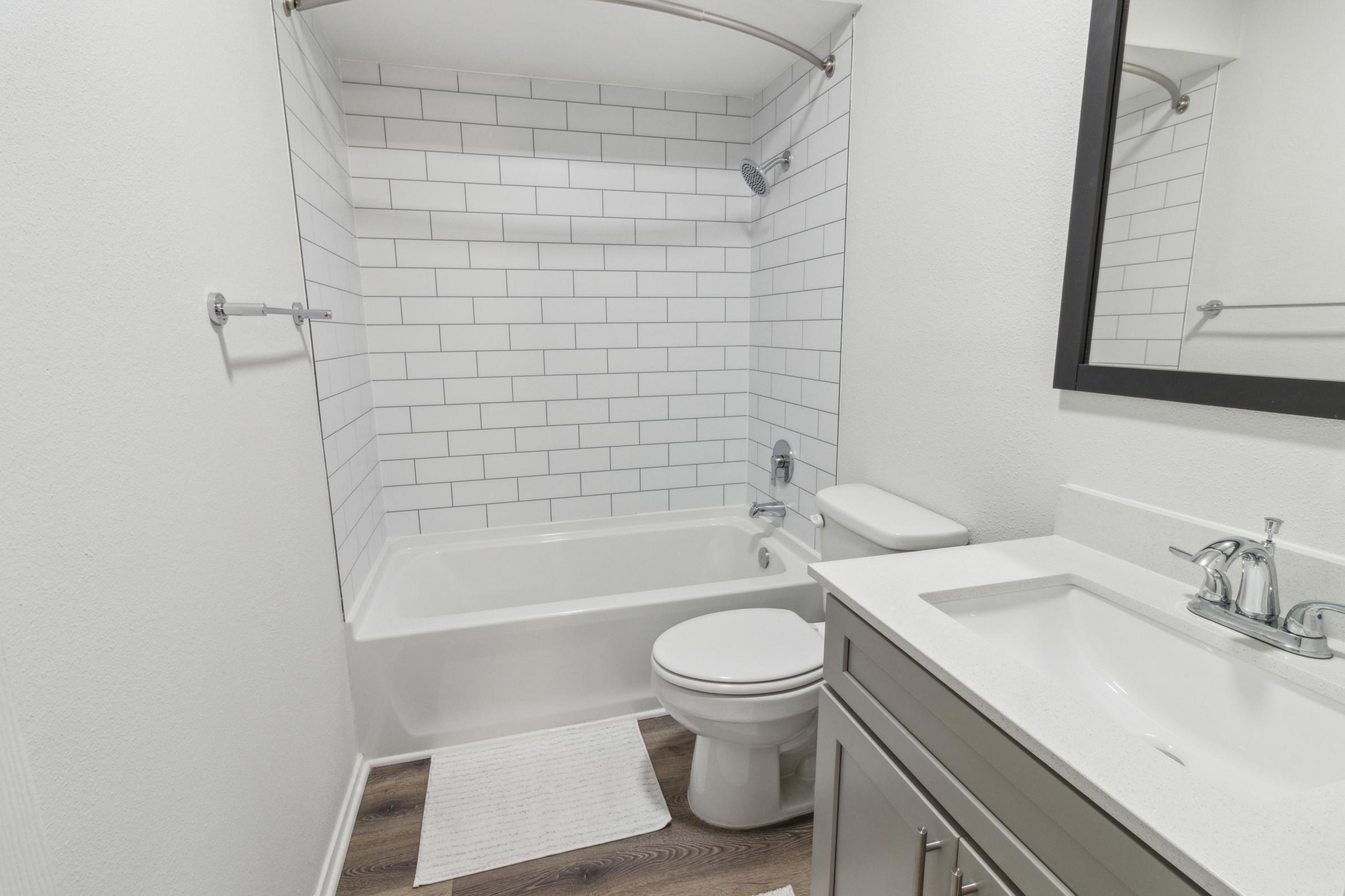 A clean, modern bathroom featuring a white tiled shower/tub combo, a white toilet, and a sleek vanity with a sink. There is a large mirror above the sink and a neutral-colored bath mat on the floor, with light-colored walls. The overall design is minimalist and contemporary.