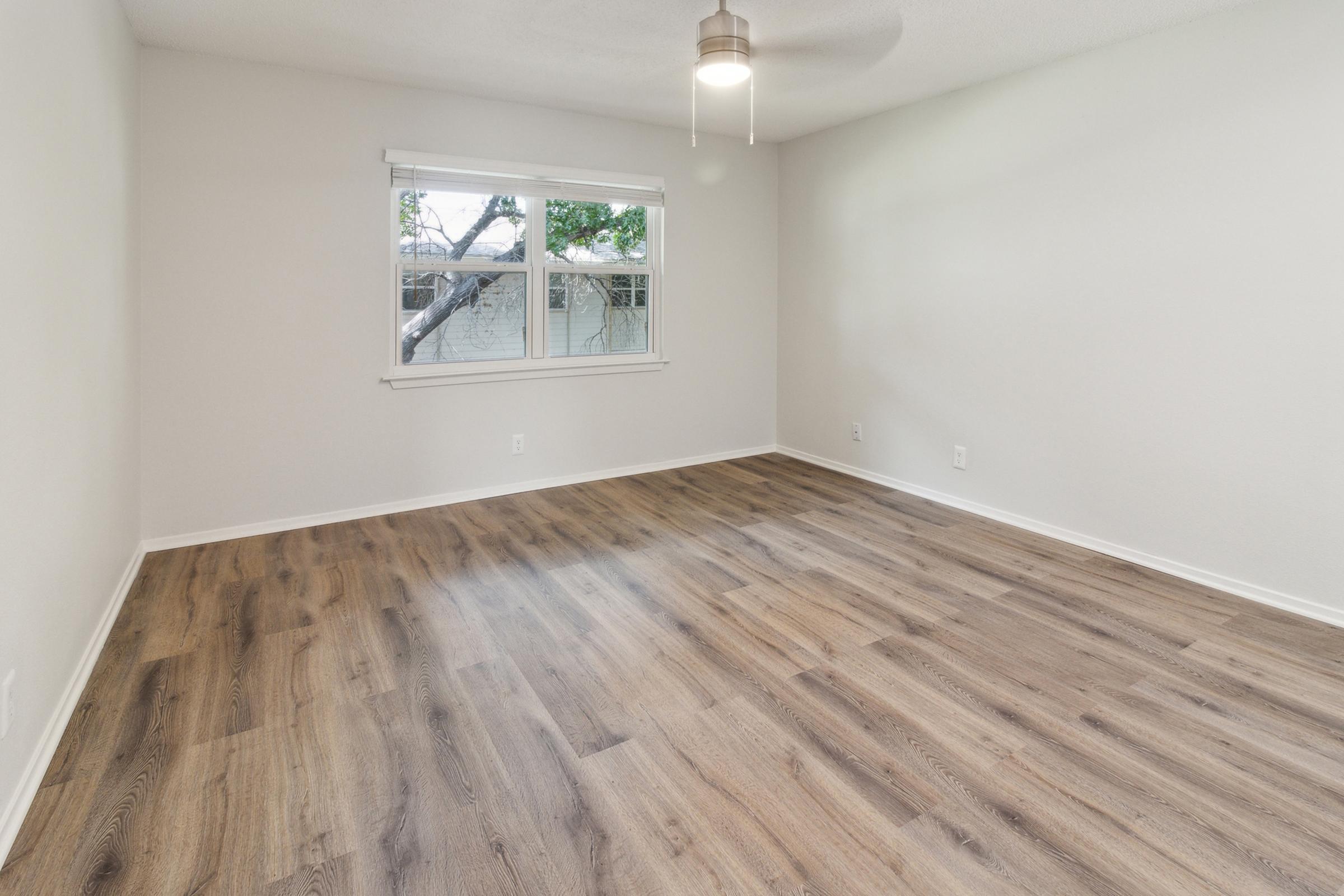 Empty room with light-colored walls and a ceiling fan. Flooring is wooden and there is a window that lets in natural light, revealing a view of trees outside. No furniture is present, creating a spacious and minimalist atmosphere.