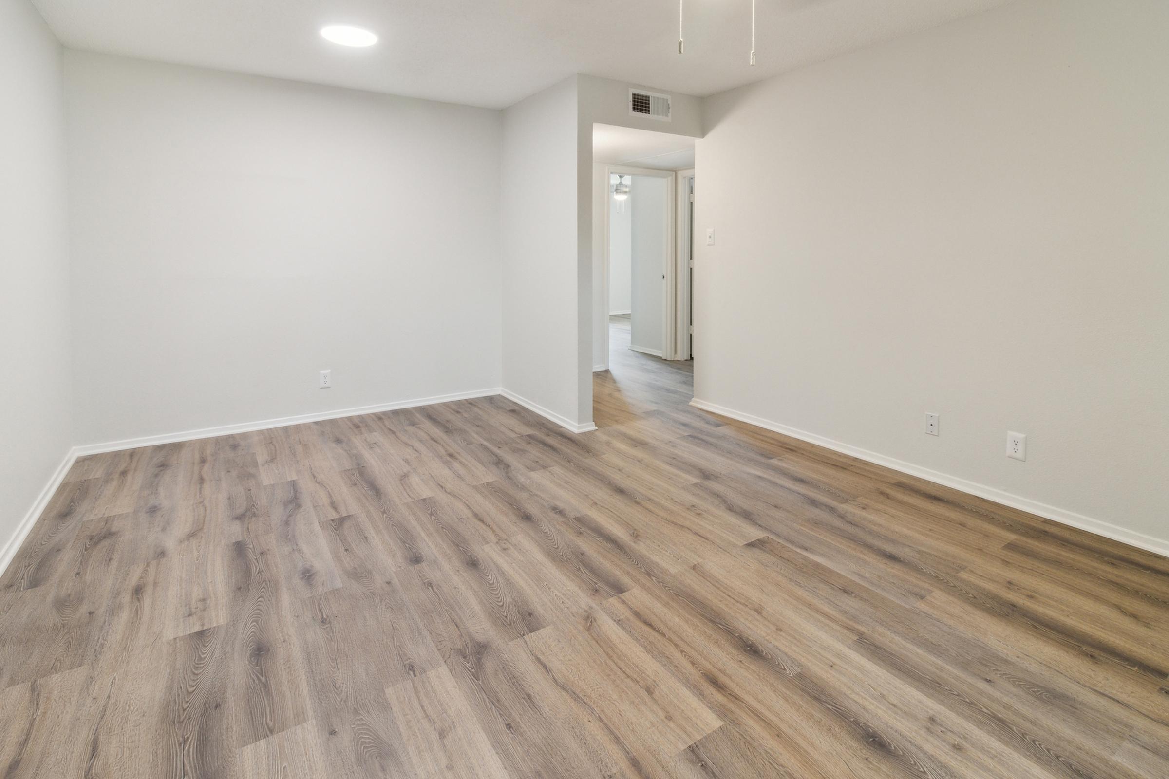 A well-lit, empty room with light-colored walls and a wood-style laminate floor. The room features a small ceiling light and an open door leading to another area, suggesting a spacious and modern living space.
