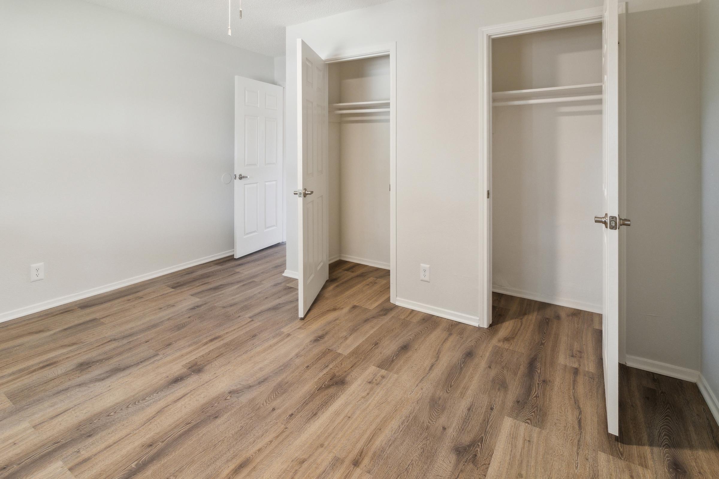 A spacious, light-colored empty room featuring two open white closet doors. The floor is covered with brown wooden planks, and the walls are painted in a neutral shade. A closed white door is visible on the left side of the image. The room has a clean and modern appearance.