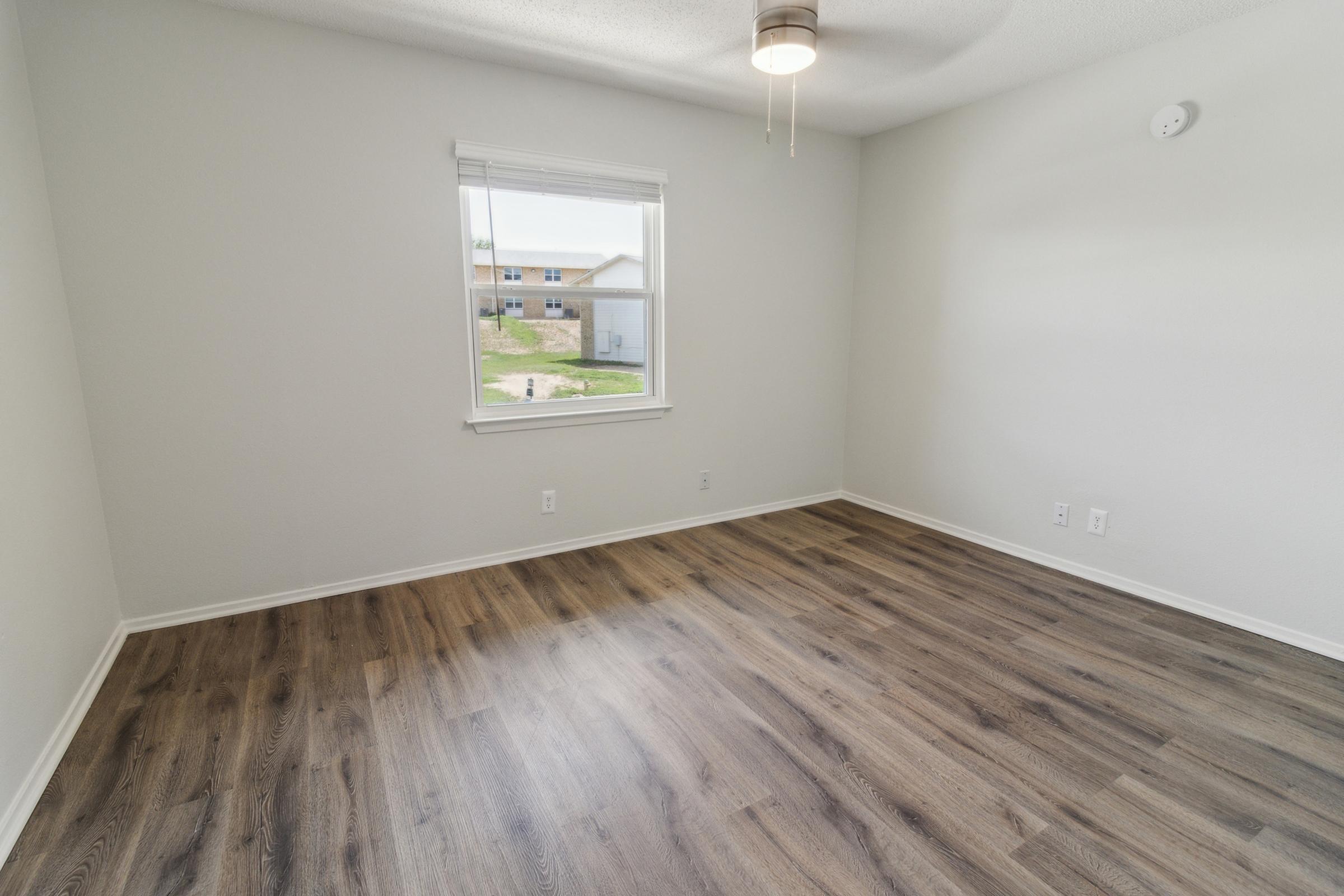 A vacant room with light-colored walls and wood-like flooring. There is a window allowing natural light, with a view of an outdoor area. A ceiling fan is installed, and there are no furnishings present, creating a spacious and airy atmosphere.