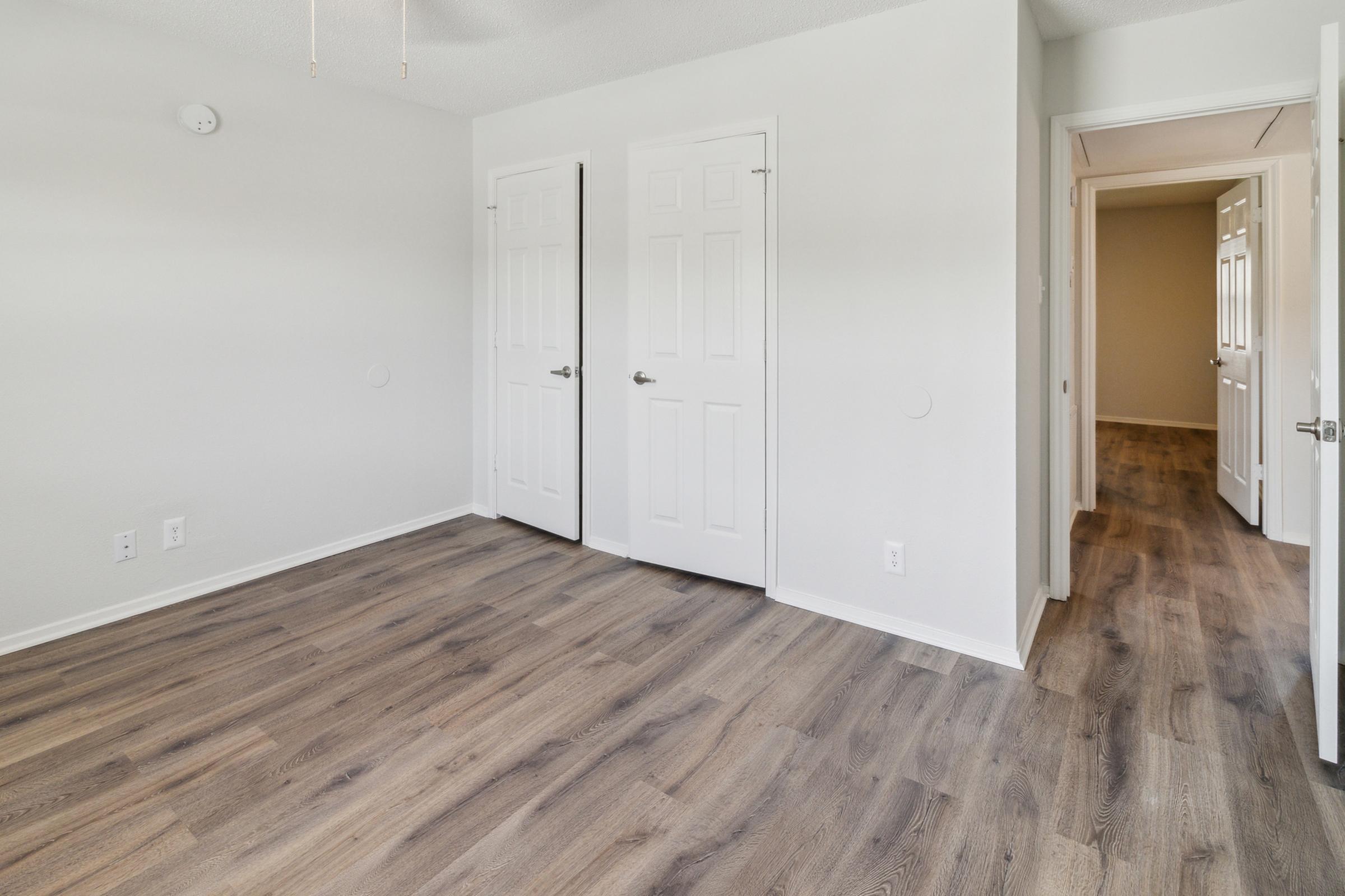A bright, unfurnished room featuring light-colored walls and wooden flooring. Two closed white doors are visible on the left side, and an open doorway is seen in the background. The ceiling has a light fixture, and the overall space appears clean and modern.