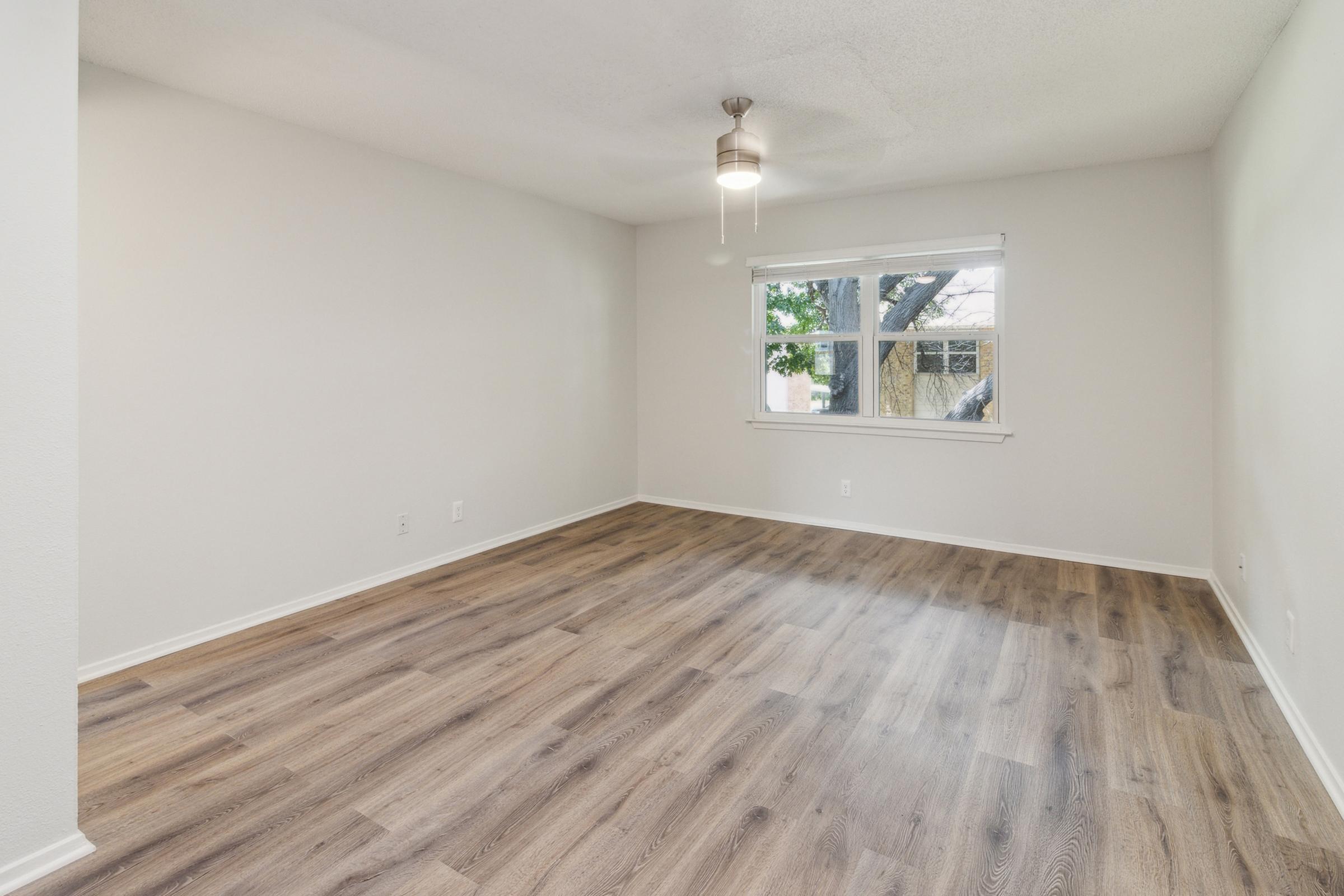 Empty room featuring light-colored walls and a smooth, wooden floor. A ceiling fan is present, and there is a large window allowing natural light to enter, framed by subtle window treatments. The room appears spacious and well-lit, suitable for various purposes.
