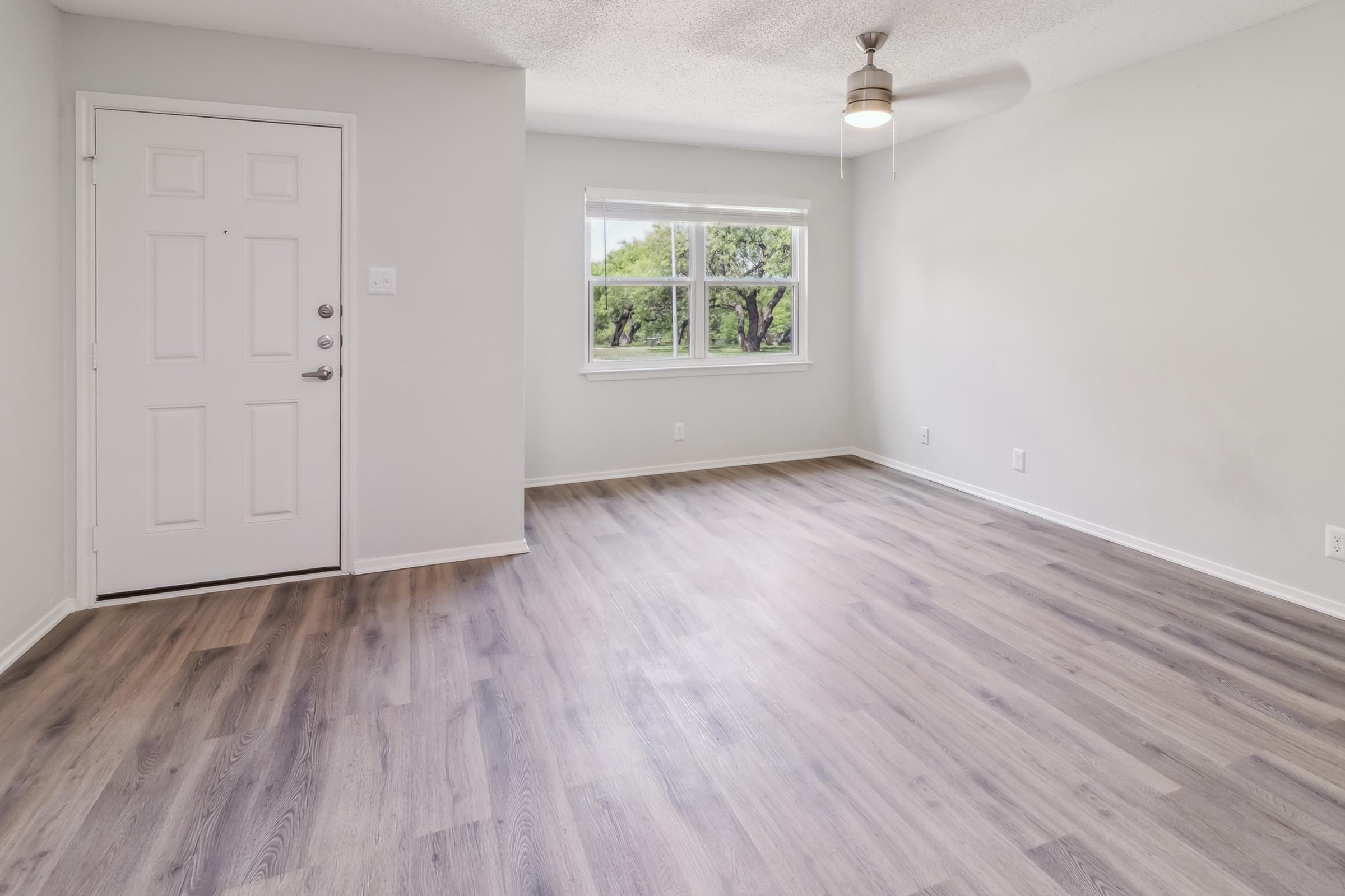 A bright, empty room featuring a light-colored wall, hardwood flooring, and a window with a view of greenery outside. The door is visible on the left side, and a ceiling fan is mounted above. The space is well-lit and has a clean, modern aesthetic.