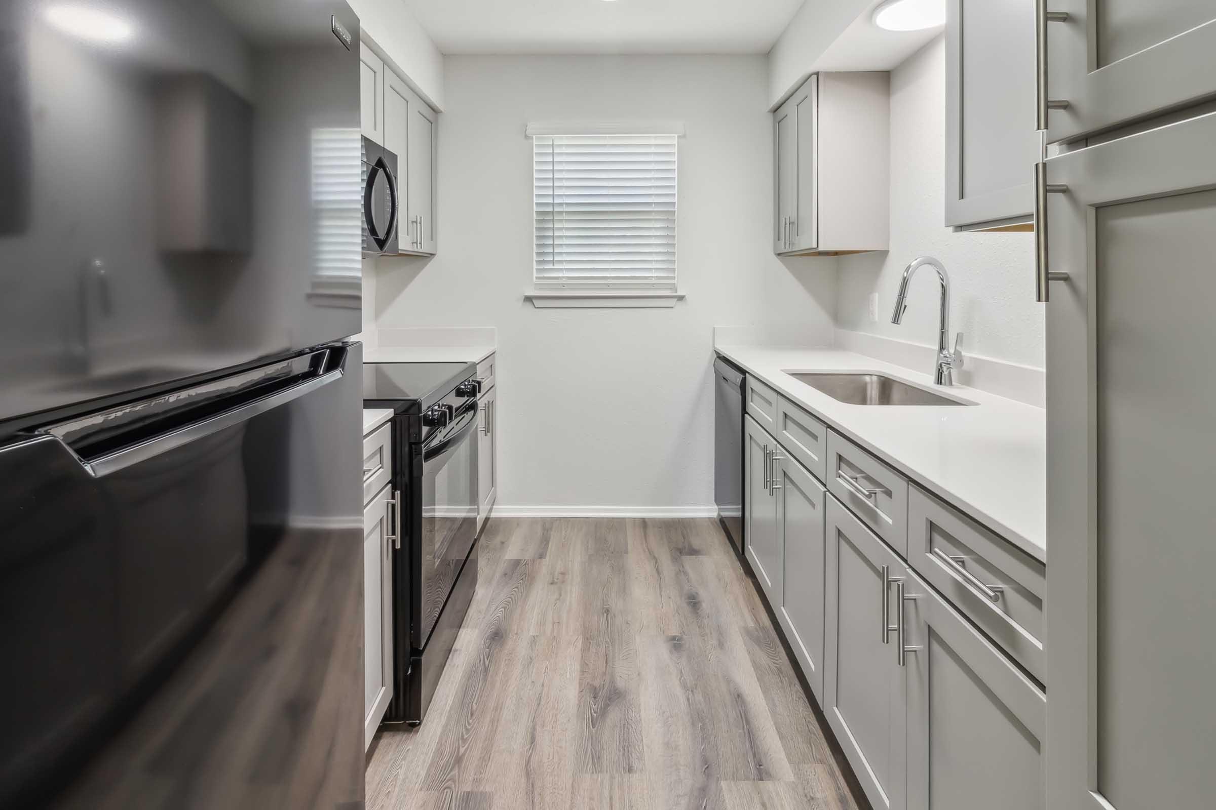 Modern kitchen featuring gray cabinets, a white countertop, a stainless steel sink, and black appliances. The space is well-lit with a window providing natural light, and has wood-like flooring that adds warmth to the minimalist design.