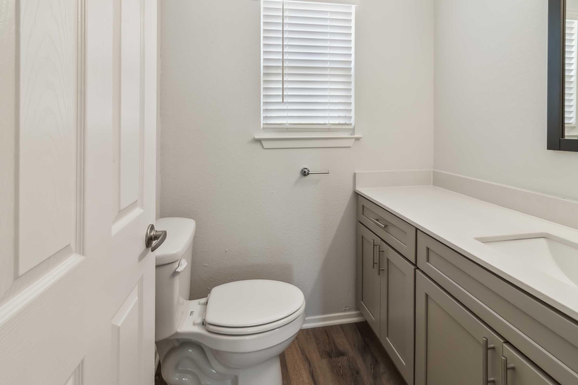 A small, modern bathroom featuring a white toilet, a light-colored vanity with cabinets, and a sink. There's a mirror above the sink and a window with blinds allowing natural light in. The walls are painted light-colored, and the flooring is a dark wood finish.