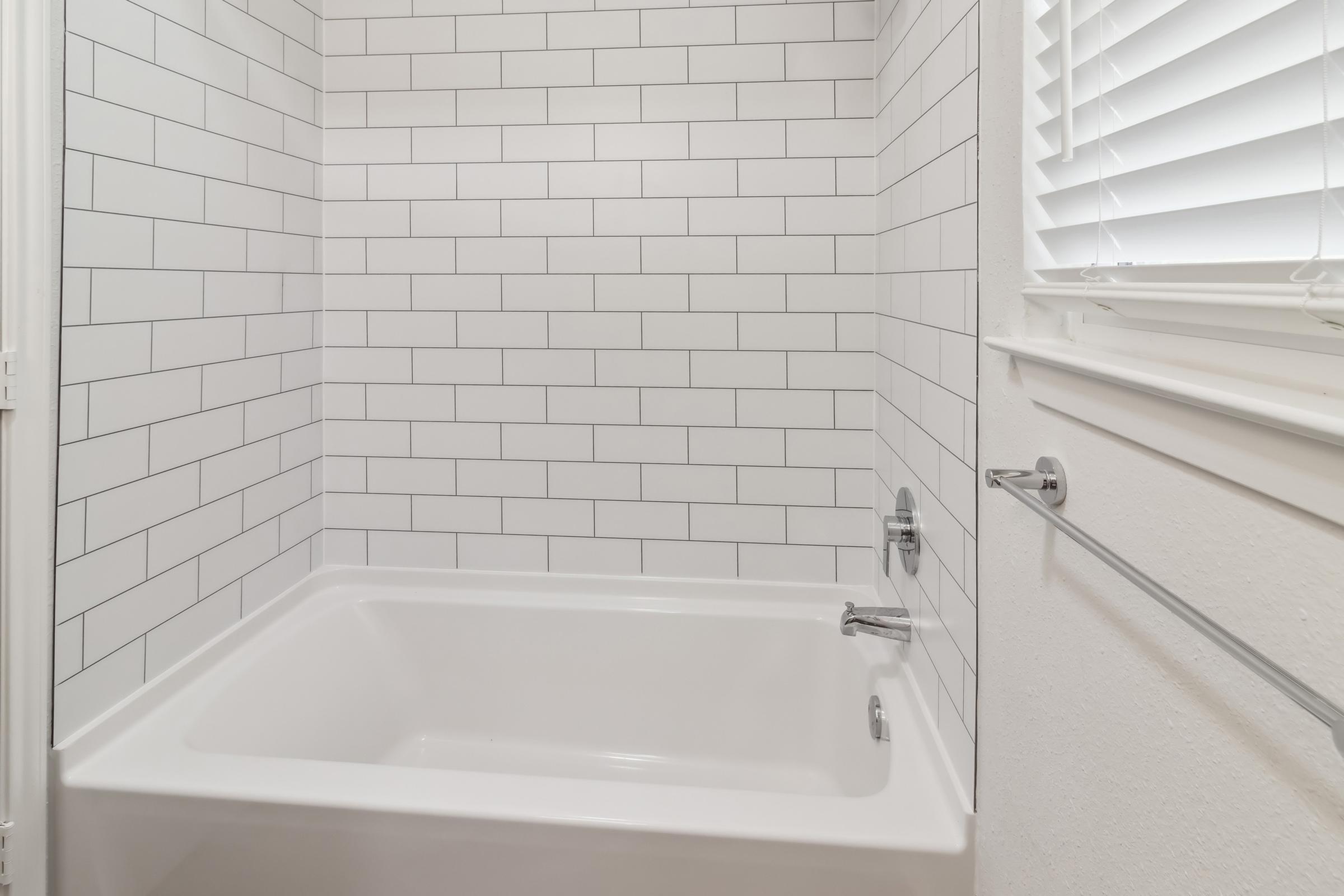 A clean, modern bathroom featuring a white bathtub with a tiled wall behind it. The tiles are white with black grout, complementing the overall bright aesthetic. A window with wooden blinds allows natural light, and there is a towel bar next to the bathtub.