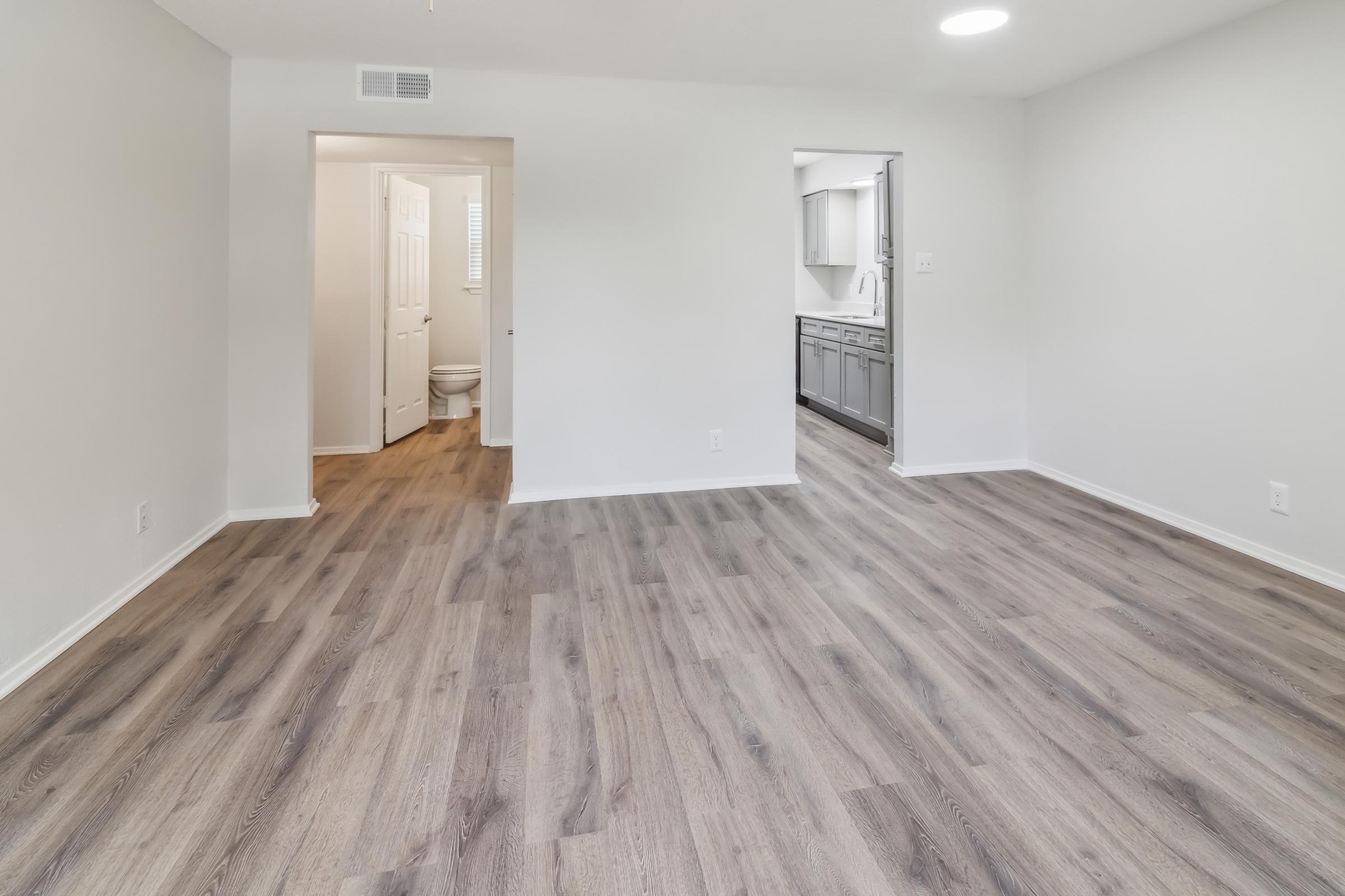 A spacious, well-lit interior of an empty apartment featuring light-colored walls and laminate flooring. The room opens to a kitchen area on one side and a bathroom door on the other, with natural light coming from a ceiling fixture. The clean, modern design offers potential for personalization.