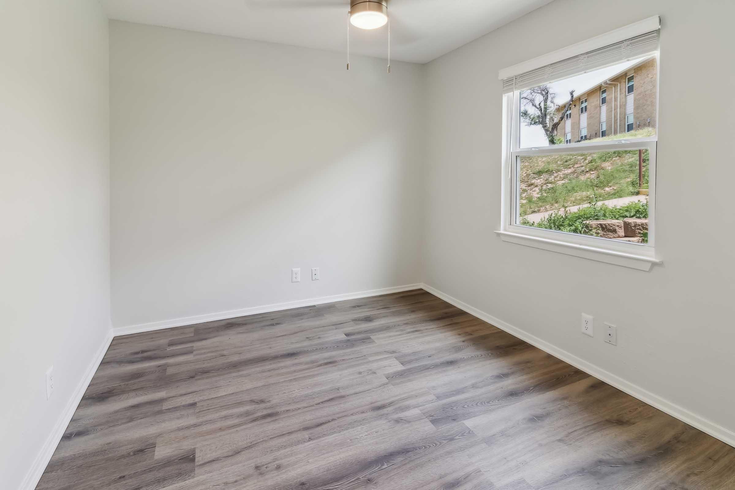 Empty room with light gray walls and a wooden floor. A ceiling fan is visible, and there's a window on the right side, showcasing a view of greenery outside. The room has a minimalist design and ample natural light.