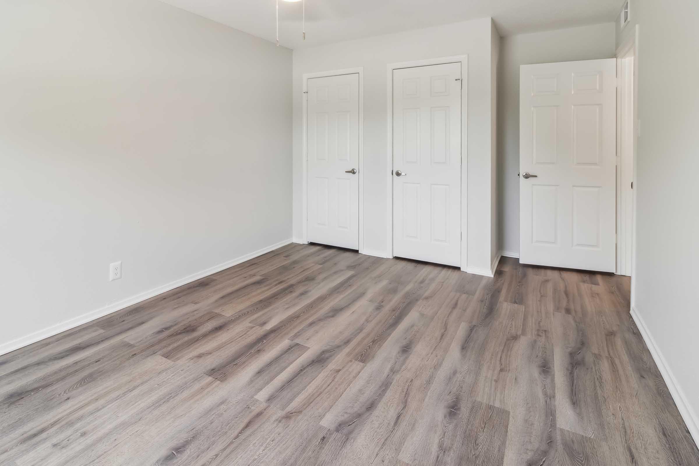 A spacious, neutral-colored room featuring light gray walls and new laminate flooring. Two closed white doors are visible, leading to closets or other areas. The room is well-lit with natural light and has a ceiling fan. The overall aesthetic is modern and minimalist, providing a clean and inviting atmosphere.