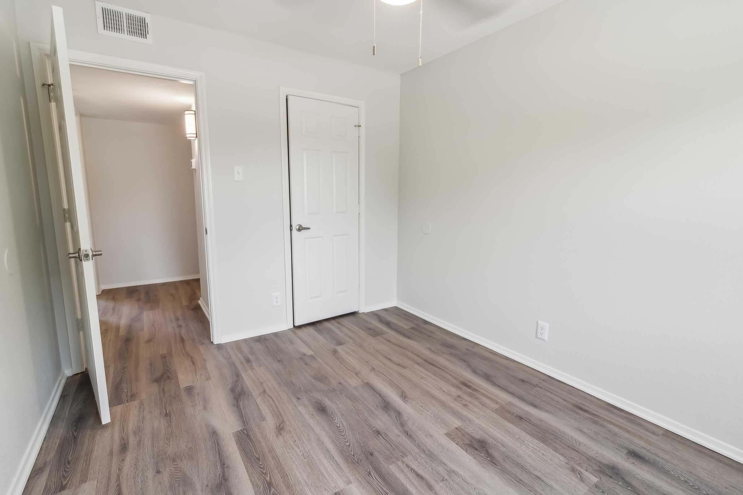 Empty room with light gray walls and a wood-like laminate floor. A door is open on the left, leading to another room, while a closed white door is on the right. The ceiling features a light fixture, and there are no furnishings in the space, giving it a clean and minimalistic appearance.