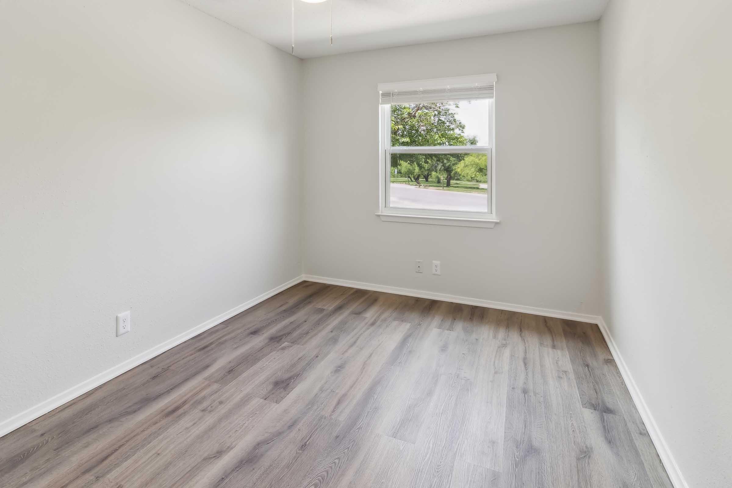 Empty room with light gray walls, a large window allowing natural light, and a view of greenery outside. The floor features light wood-like laminate. The space is uncluttered and ideal for various uses, such as an office or bedroom.