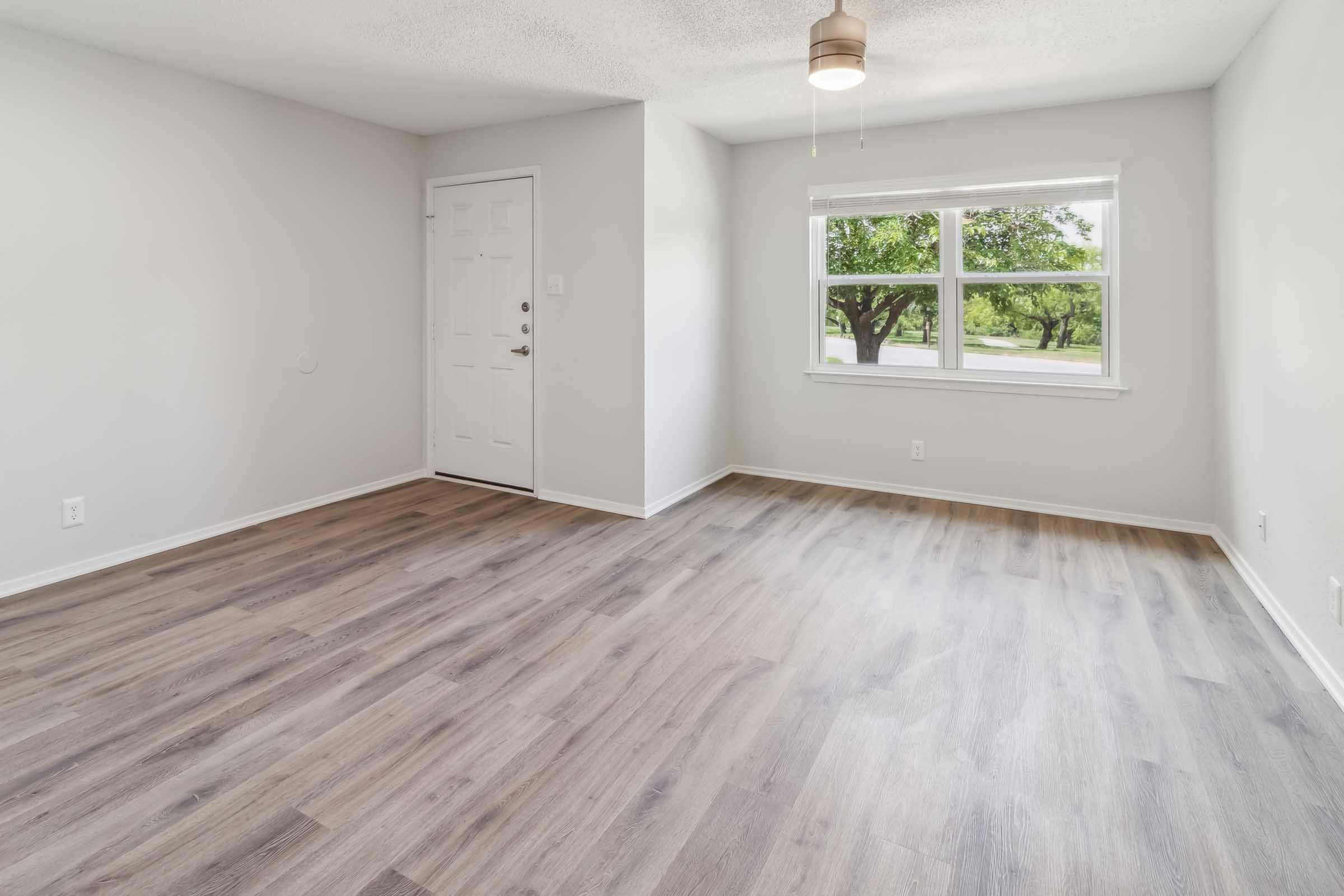 A well-lit, empty room with light gray walls and a ceiling fan. The floor features light wood-like laminate. There's a window with views of greenery and a front door on the left side. The space is clean and modern, suitable for various uses.