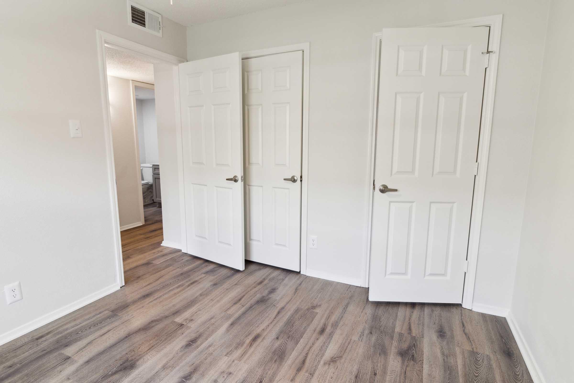 A clean, empty room featuring light gray walls and a wood-like floor. There are three white doors on the right side, two of which are slightly ajar, leading to different spaces. Soft natural light enters from the doorway at the back, creating a spacious and bright atmosphere.