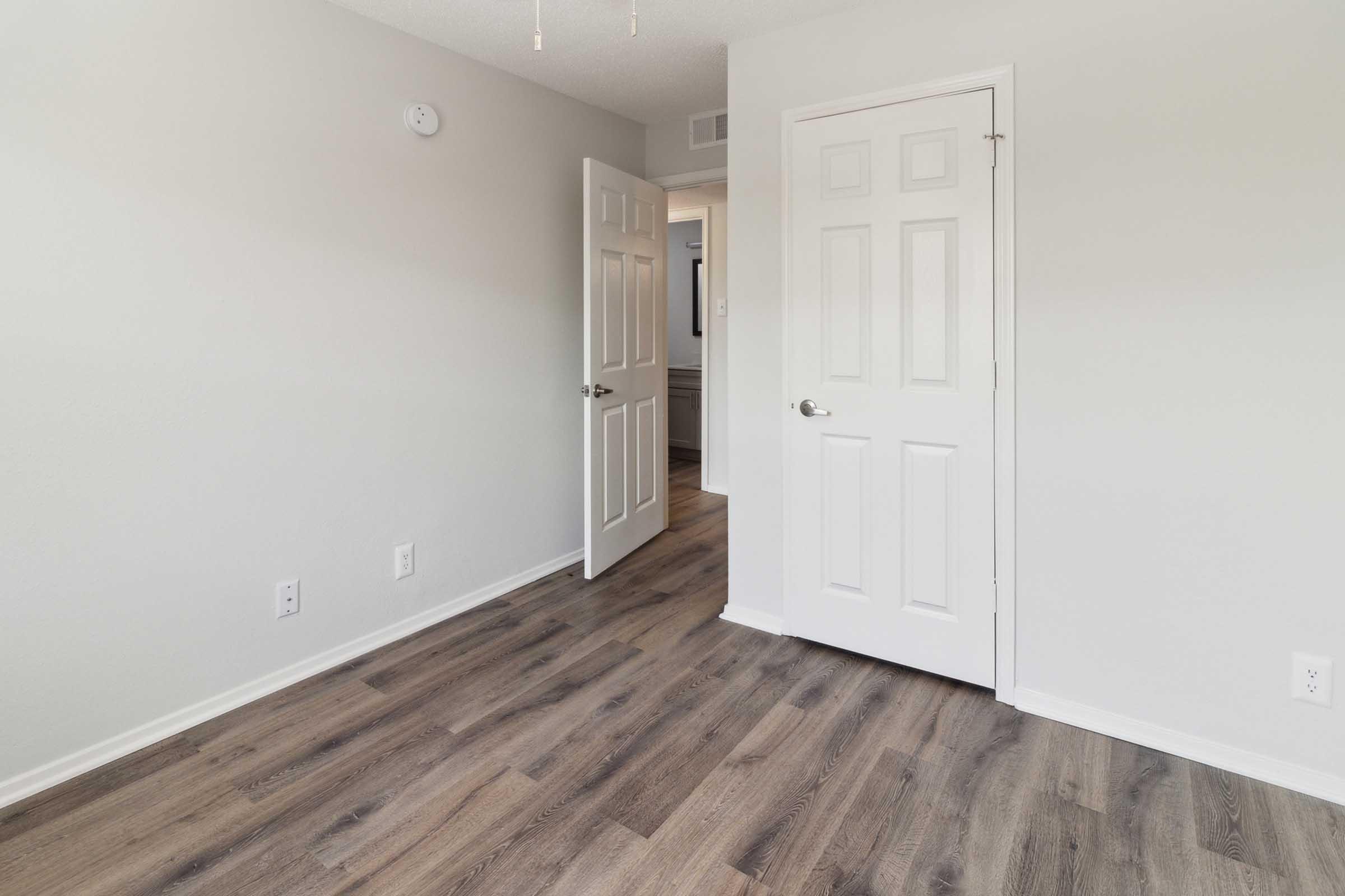 A view of a light-colored room featuring two closed doors on the left and a wall-mounted smoke detector. The floor is made of laminate with wood-like texture, and the walls are painted in a soft neutral shade. The room appears clean and well-lit, suitable for various uses.