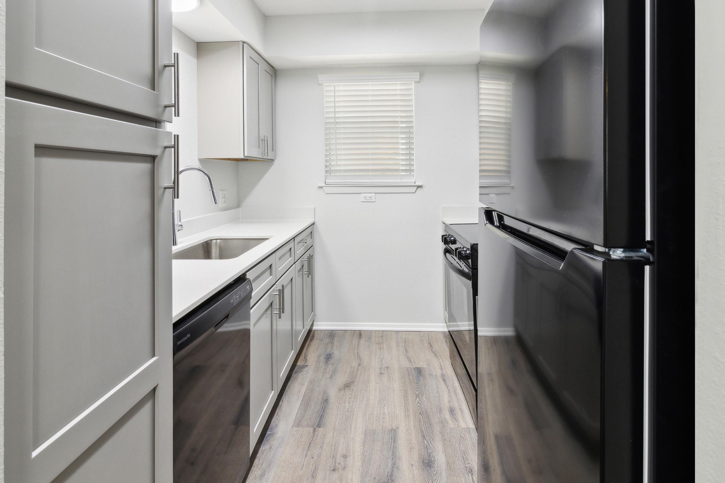 Modern kitchen featuring stainless steel appliances, including a dishwasher and a black refrigerator. The cabinets are light-colored, and a window with white blinds allows natural light to enter. The flooring is wood-like laminate, creating a warm and inviting atmosphere.