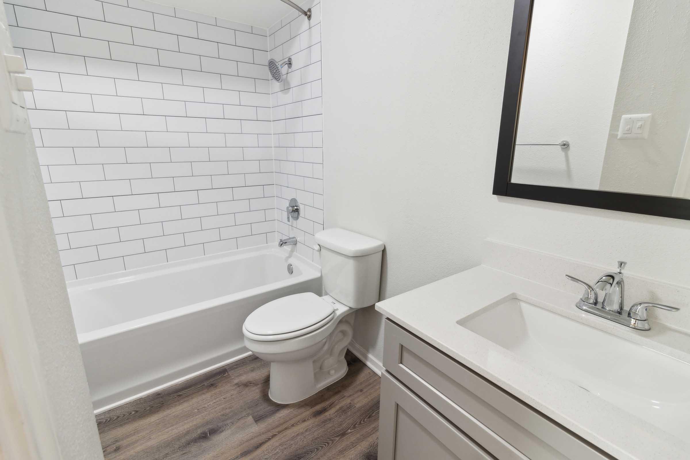 A modern bathroom featuring a white bathtub with a shower, a white toilet, and a vanity with a sink. The walls are adorned with white subway tiles, and there is a large mirror above the sink. The floor is made of laminate wood, adding warmth to the space.