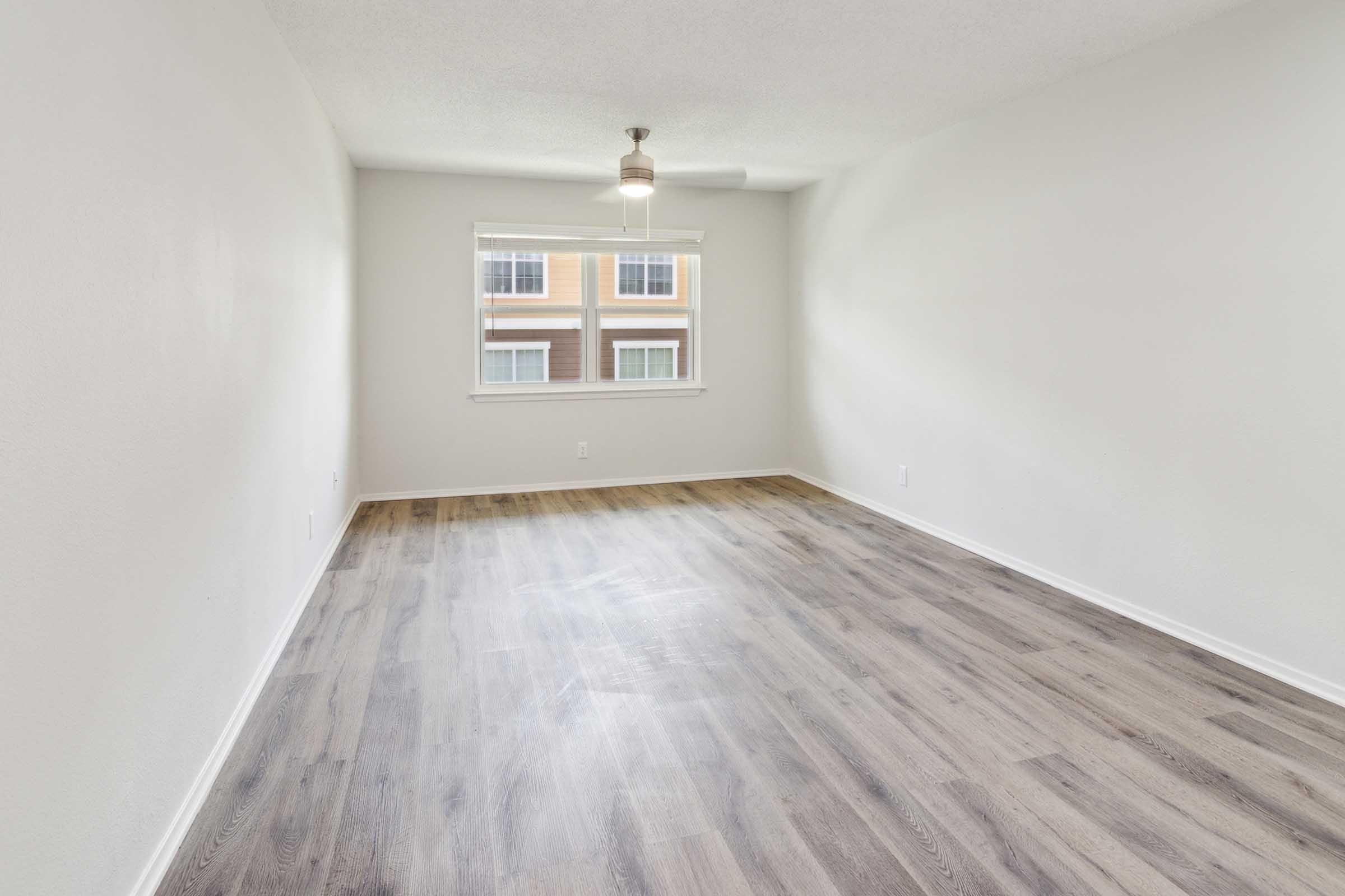 A spacious empty room with light-colored walls and a ceiling fan. The floor is covered in a light wood laminate. A window allows natural light to enter, revealing the view of another building outside. The room is uncluttered, providing a blank canvas for future decoration.