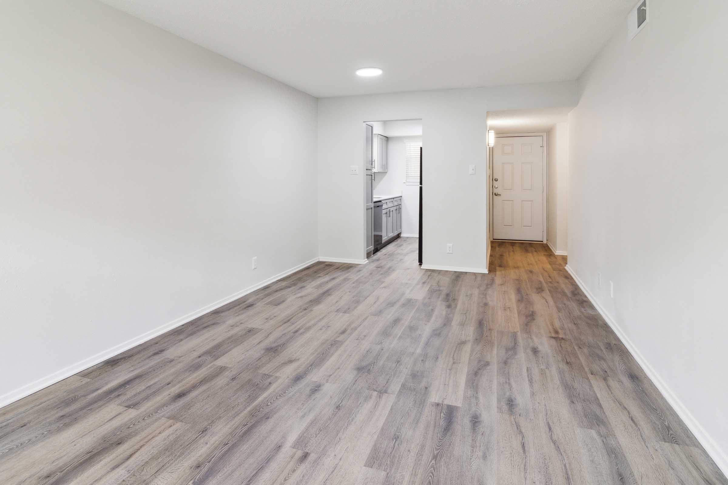 A spacious, empty room featuring light-colored walls and laminate flooring. One side has a doorway leading to a kitchen area, while another doorway leads to an entrance. Ample natural light is present, enhancing the bright and airy atmosphere of the space.
