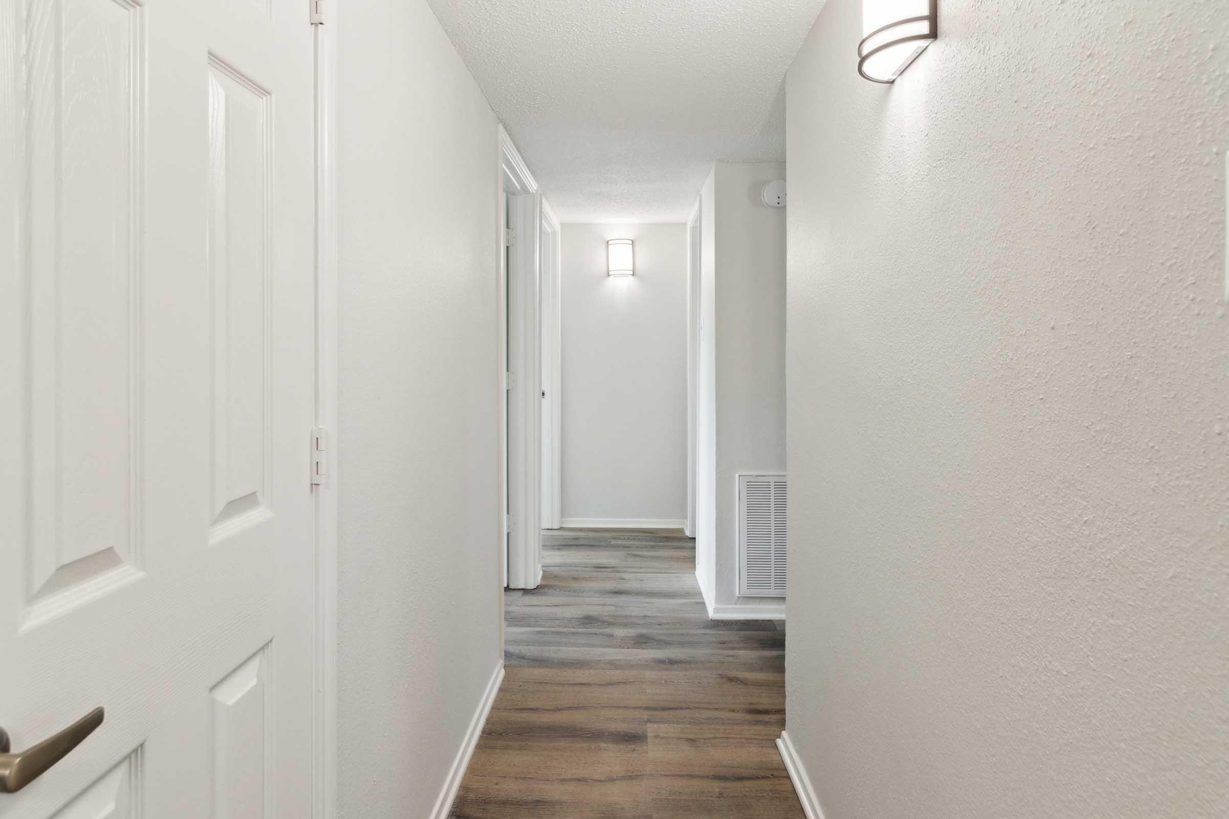 A well-lit hallway with light gray walls and modern wall sconces. The floor features light-colored wooden planks. Doorways are visible on either side, leading to other rooms, and there is a door at the end of the hallway, contributing to a clean and spacious look.