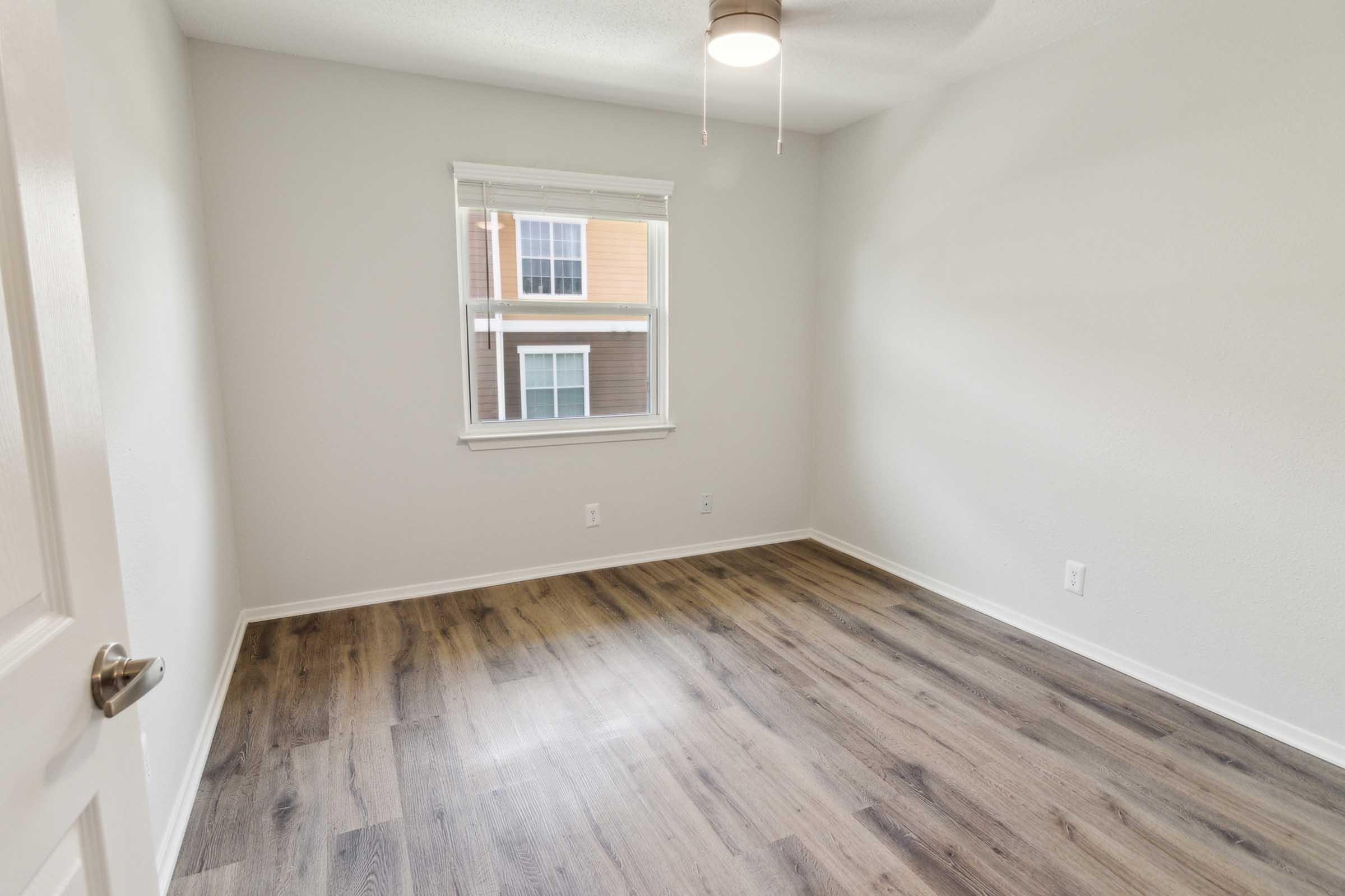 A vacant room with light-colored walls and a ceiling fan. The floor is made of wood laminate, and there is a window allowing natural light to enter. The view outside the window shows the exterior of a building. The space appears clean and ready for furniture placement.