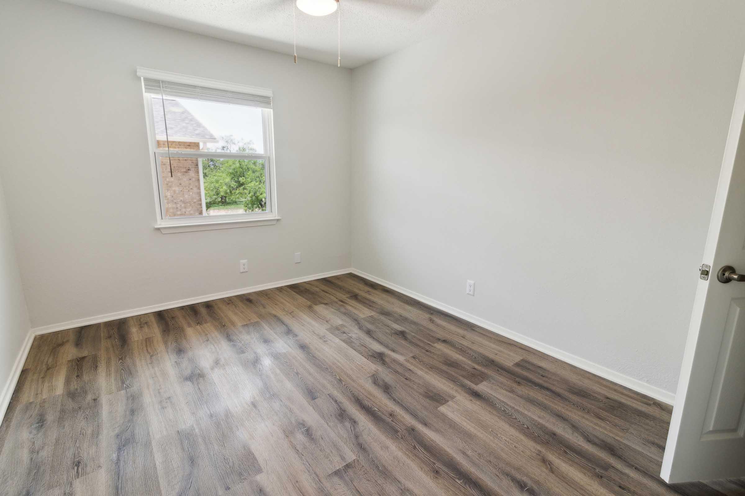 A clean, empty room with light gray walls and a medium-toned wooden floor. The room has a window letting in natural light and a ceiling fan. There are no furnishings or decorations. The door is closed, and the space appears well-maintained and ready for use.