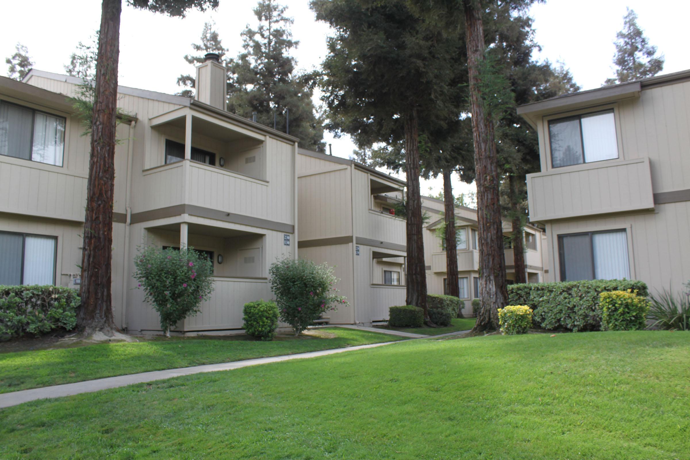 Two-story residential apartment complex surrounded by green lawns and trees. The buildings feature balconies and windows, with landscaped shrubs and plants in the foreground. The scene conveys a quiet, suburban atmosphere with well-maintained outdoor spaces.