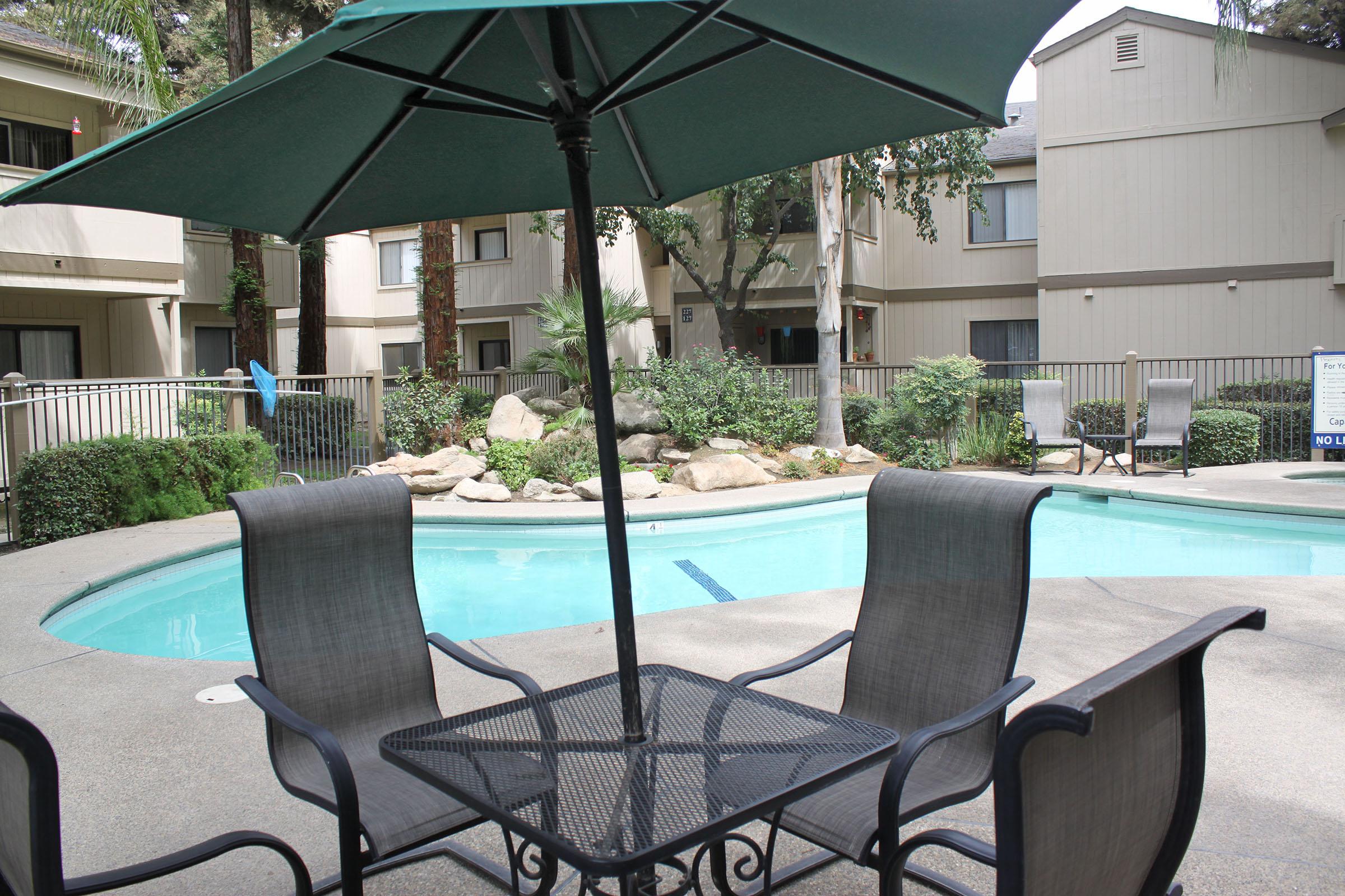 A cozy outdoor pool area featuring a circular swimming pool surrounded by greenery and rocks. In the foreground, there is a black metal table with four chairs and a green umbrella, providing shade. Apartment buildings can be seen in the background, adding to the serene atmosphere.
