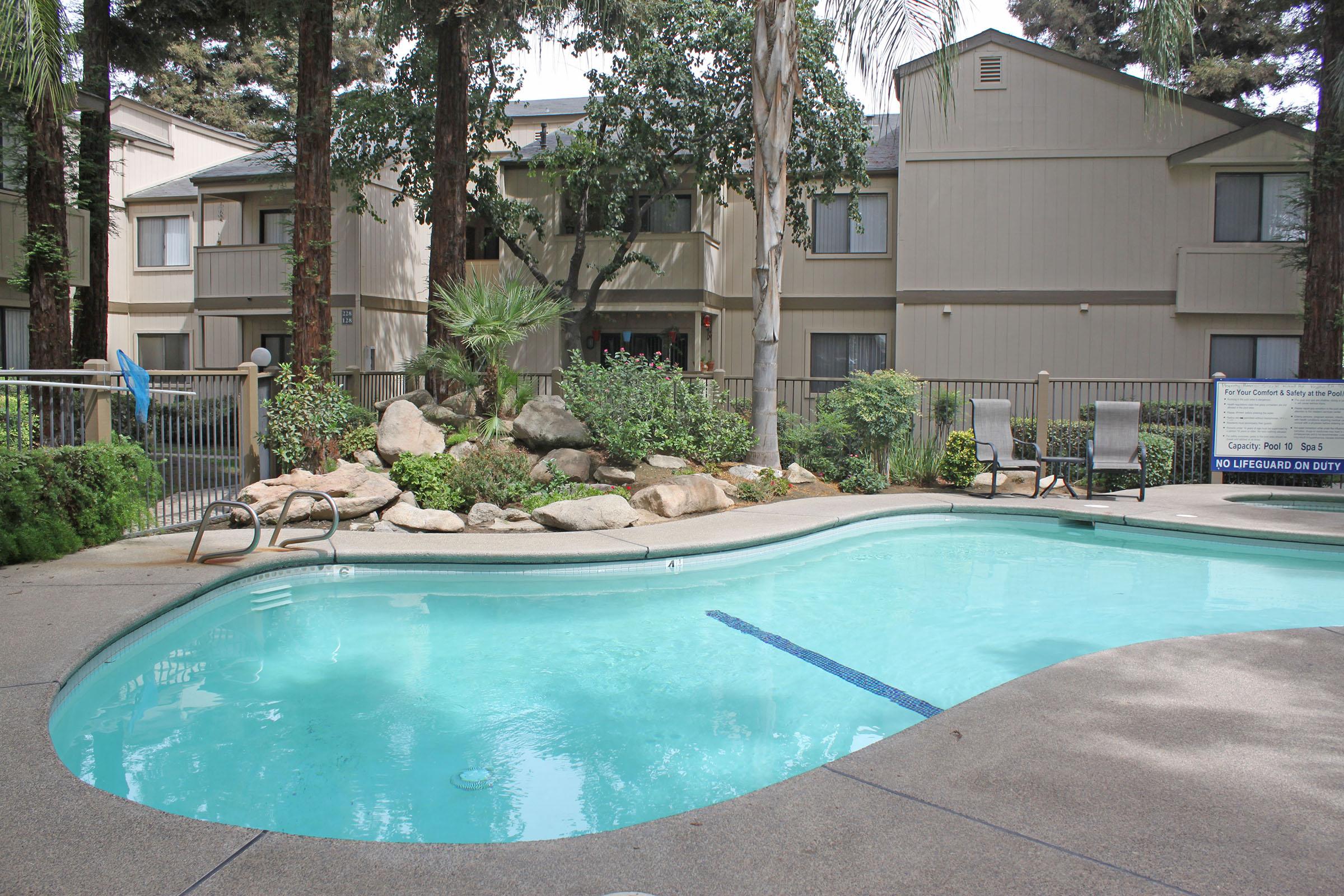 A clear swimming pool surrounded by shrubs and rocks, with lounge chairs on the concrete deck. In the background, there are apartment buildings framed by trees, creating a peaceful outdoor setting. A sign near the pool emphasizes safety and caution.
