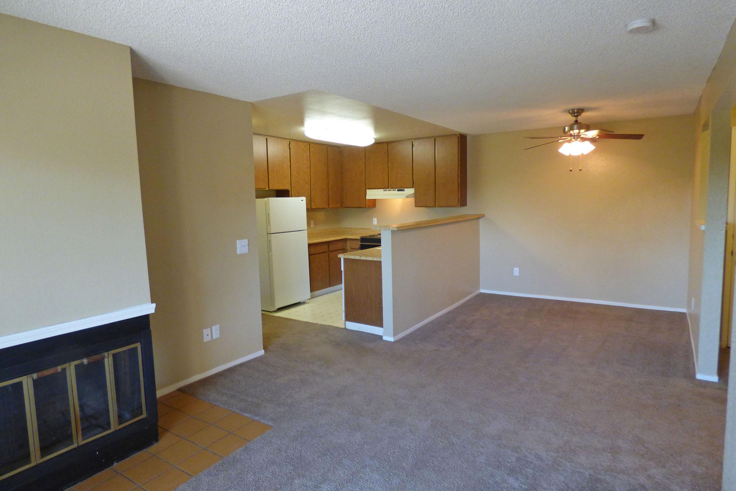 A spacious living area with neutral-colored walls and carpet. The open layout includes a kitchen with wooden cabinets, a white refrigerator, and a ceiling fan. A fireplace is visible on the left side of the room, enhancing the cozy atmosphere. Bright natural light illuminates the space.