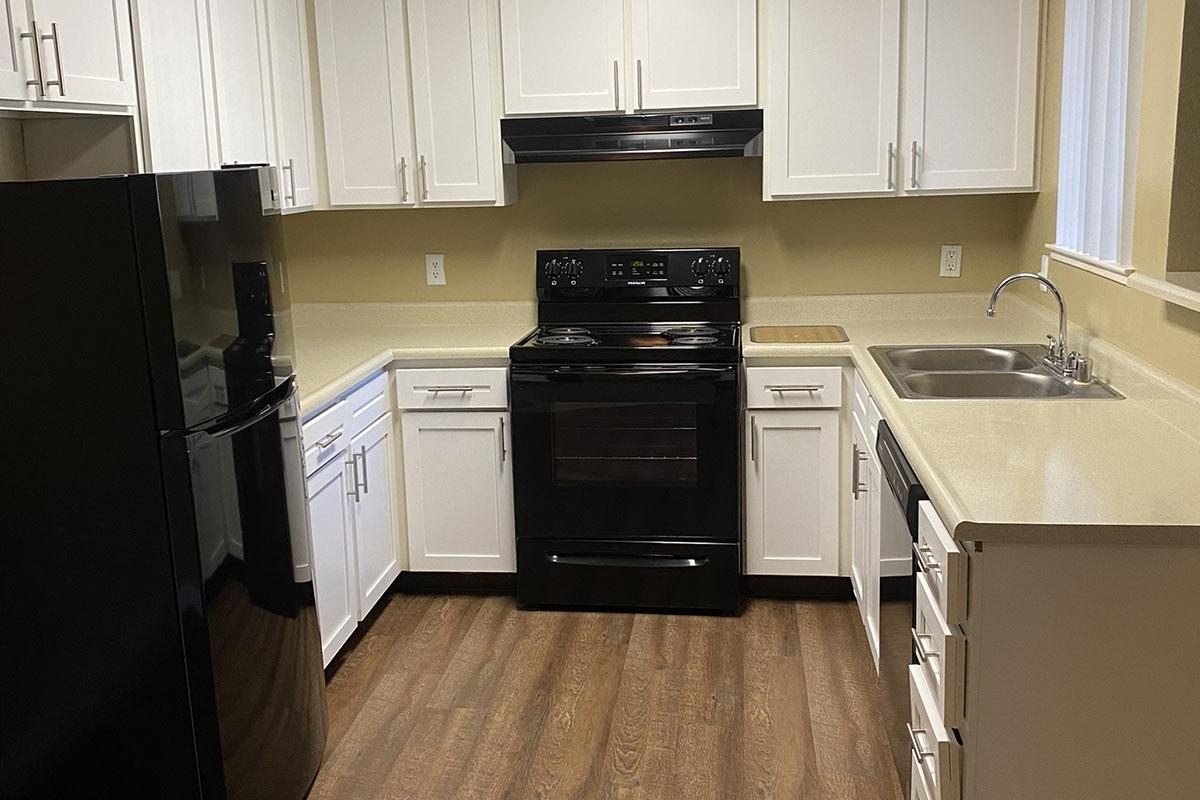 A modern kitchen featuring white cabinets, a black refrigerator, and a black oven/stove. The countertops are light-colored, and there is a stainless steel sink next to the stove. The flooring is wood-like, giving the space a warm and inviting appearance. Natural light comes from a nearby window.