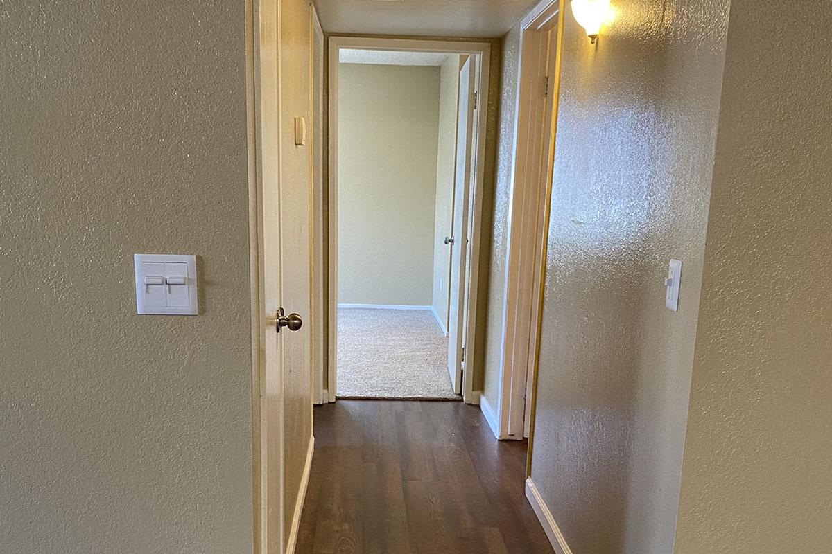 A narrow hallway with light-colored walls and a wooden floor. There are two doorways at the end, one leading to a carpeted room and the other potentially leading outside the frame. A light switch is visible on the left wall, and warm lighting creates a welcoming atmosphere.
