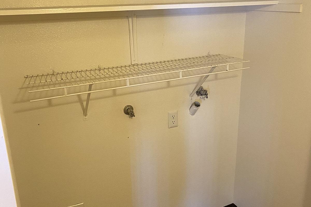 Empty closet space with a white wire shelf mounted on a beige wall. There are two utility fixtures on the wall: a water supply valve and a drainage pipe. The floor is bare, and the overall area appears tidy and ready for storage or organization.
