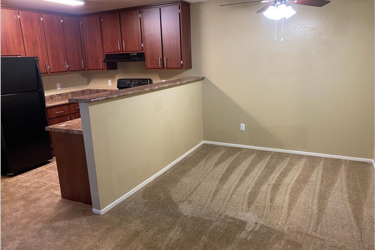 A spacious kitchen area with dark wood cabinets, a black refrigerator, and a countertop overlooking a beige-carpeted living space. The walls are painted a light green shade, and there is a ceiling fan. Natural light creates shadows on the carpeted floor, highlighting the open design of the room.
