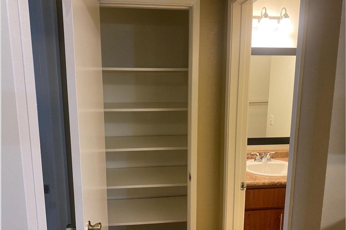 A well-lit closet with empty shelves on the left, leading to a small bathroom on the right. The bathroom has a double sink with a countertop, a mirror above it, and bright overhead lights. The walls are painted in neutral colors, creating a clean and organized appearance.