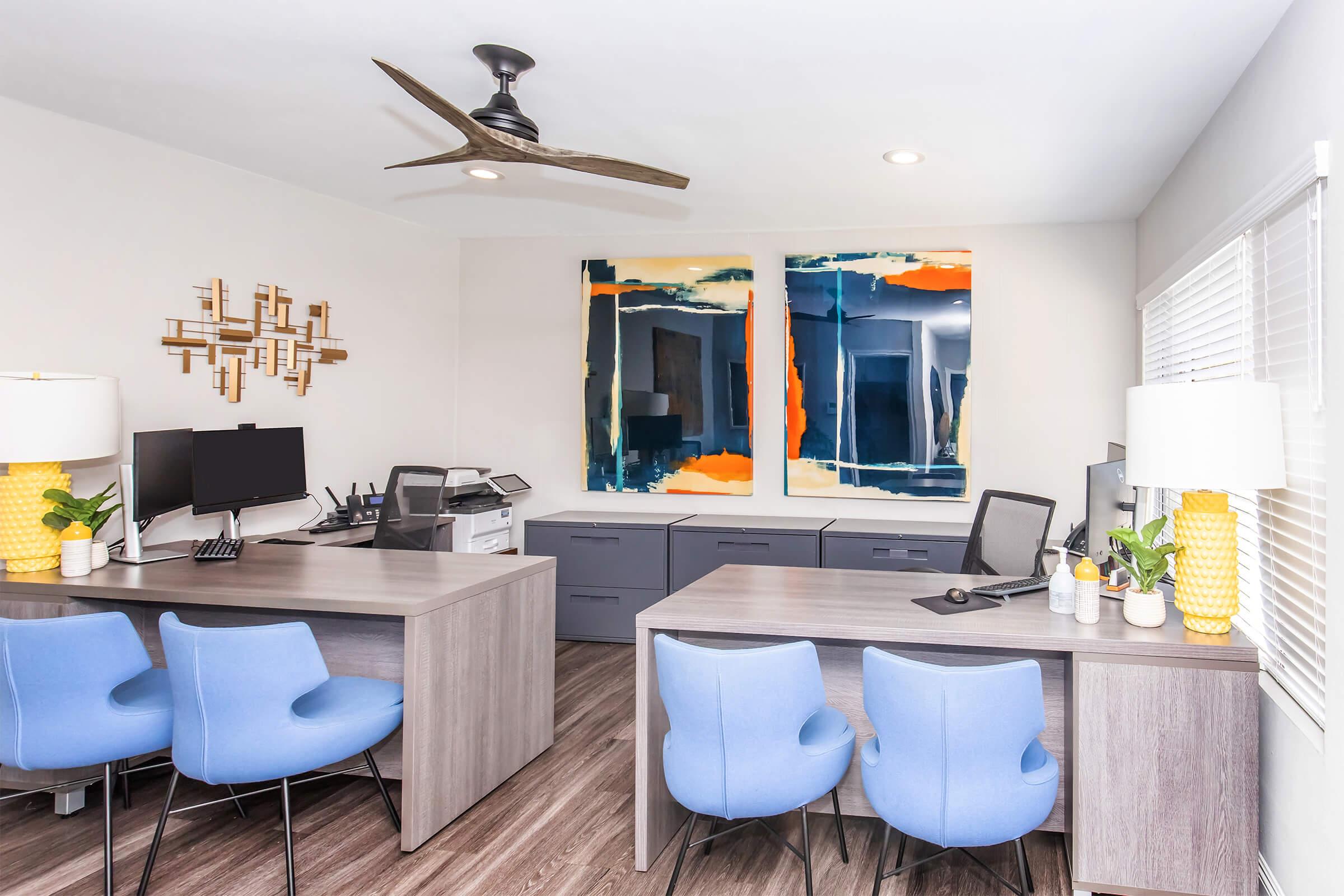 A modern office space featuring two wooden desks with blue chairs, a printer, and dual computer screens. The walls are adorned with abstract artwork in vibrant colors. A ceiling fan is visible, and natural light streams in through window blinds, creating a bright and inviting atmosphere.