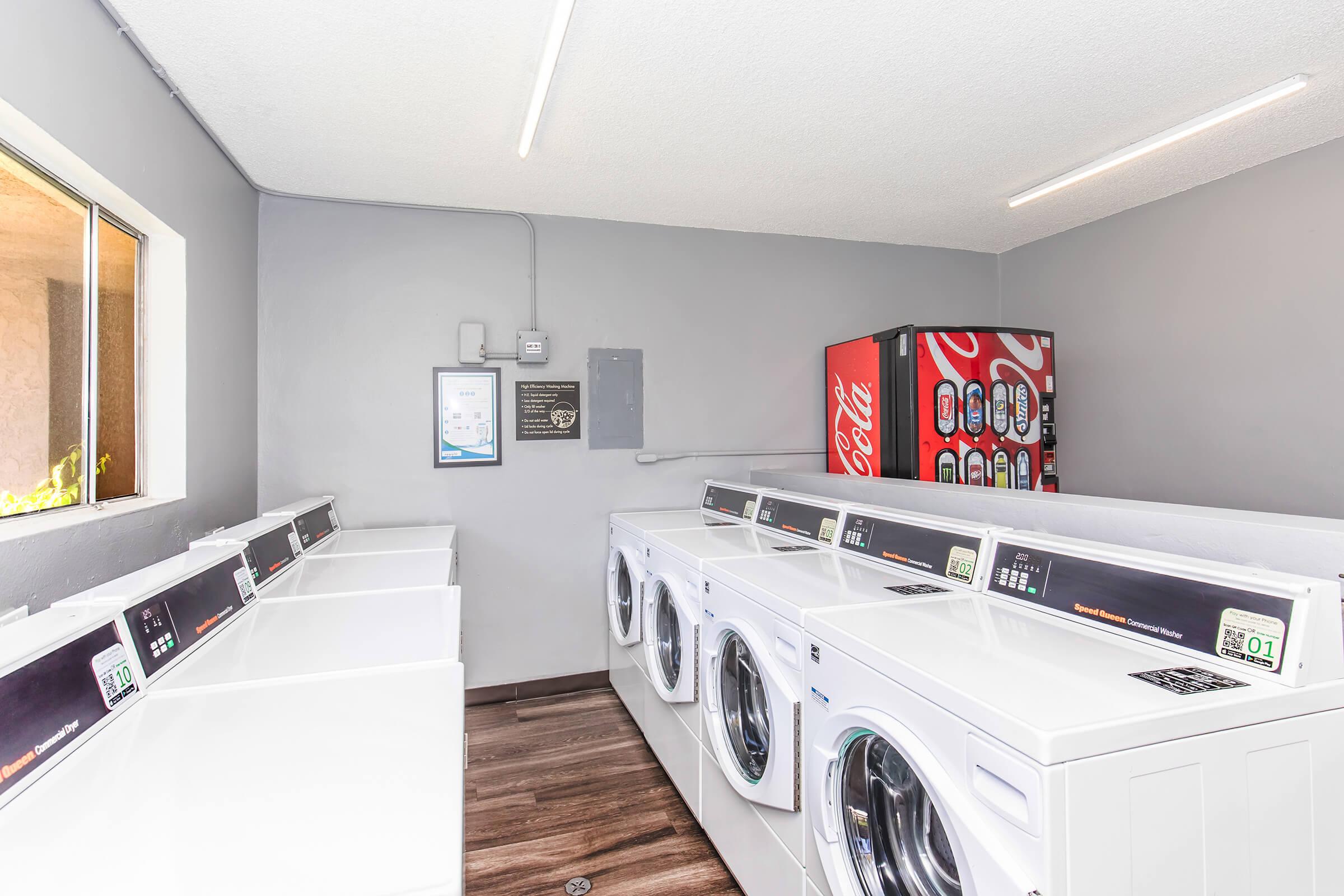 A modern laundry room featuring several white washing machines and a vending machine stocked with Coca-Cola products. The walls are painted gray, and there is bright overhead lighting and a window allowing natural light in. The flooring is wooden.